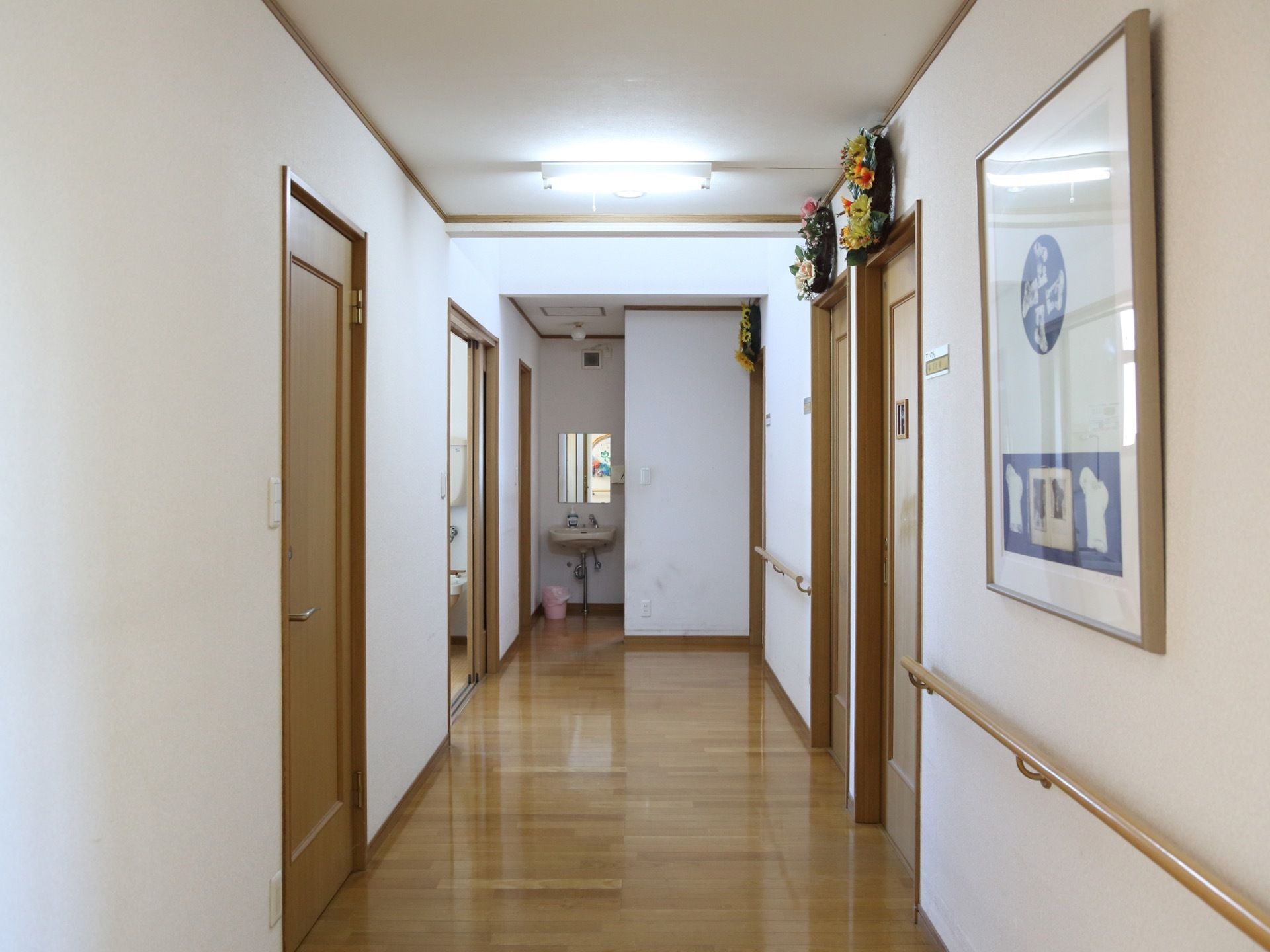 通路の片側に手すりが設置されている。扉の上には花が飾られている。通路の奥に手洗い場があるのが見える。