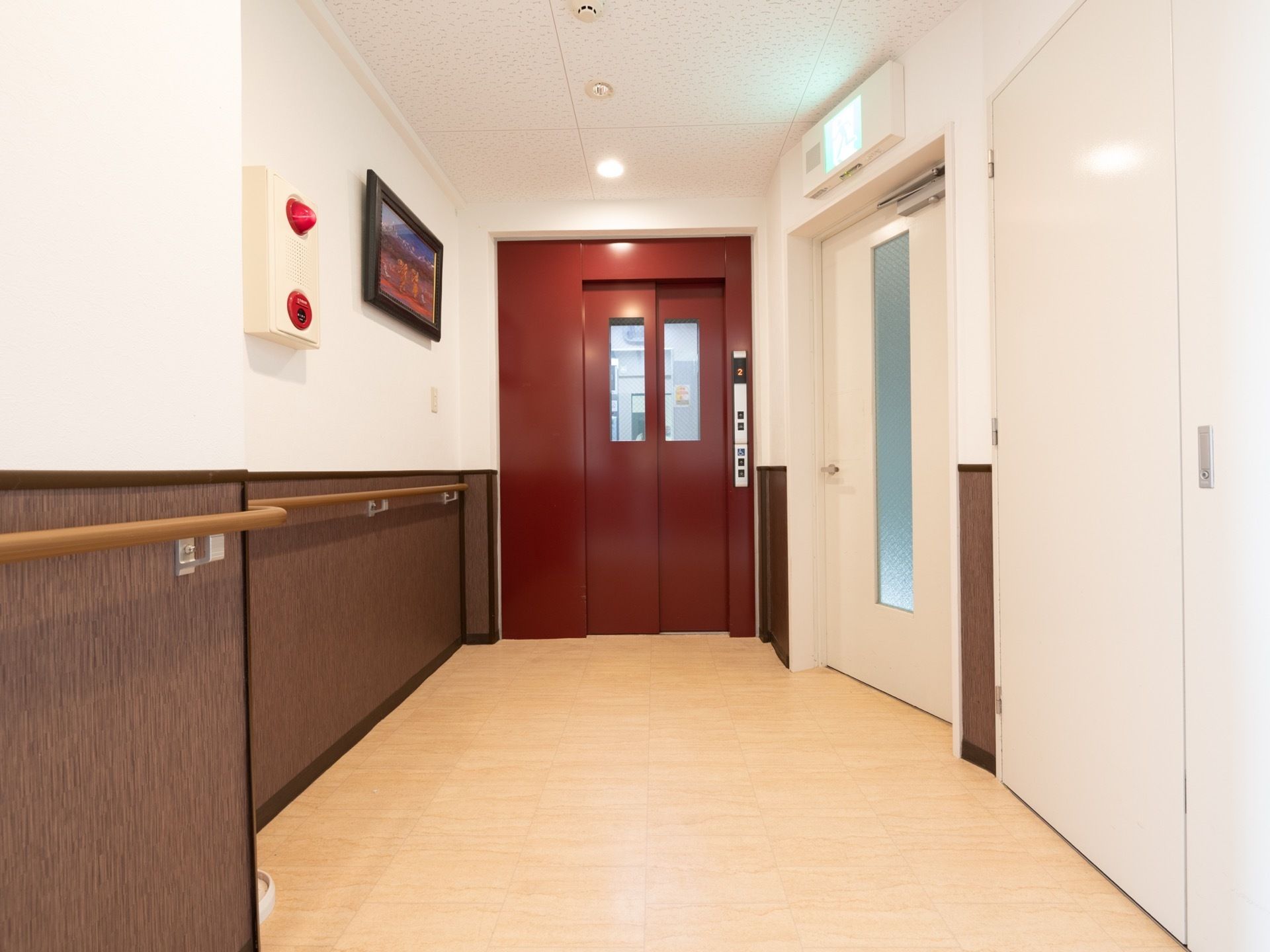 通路の突き当りにエレベーターがある。右側には避難口、反対側の壁には火災報知器があり、絵画が飾られている。
