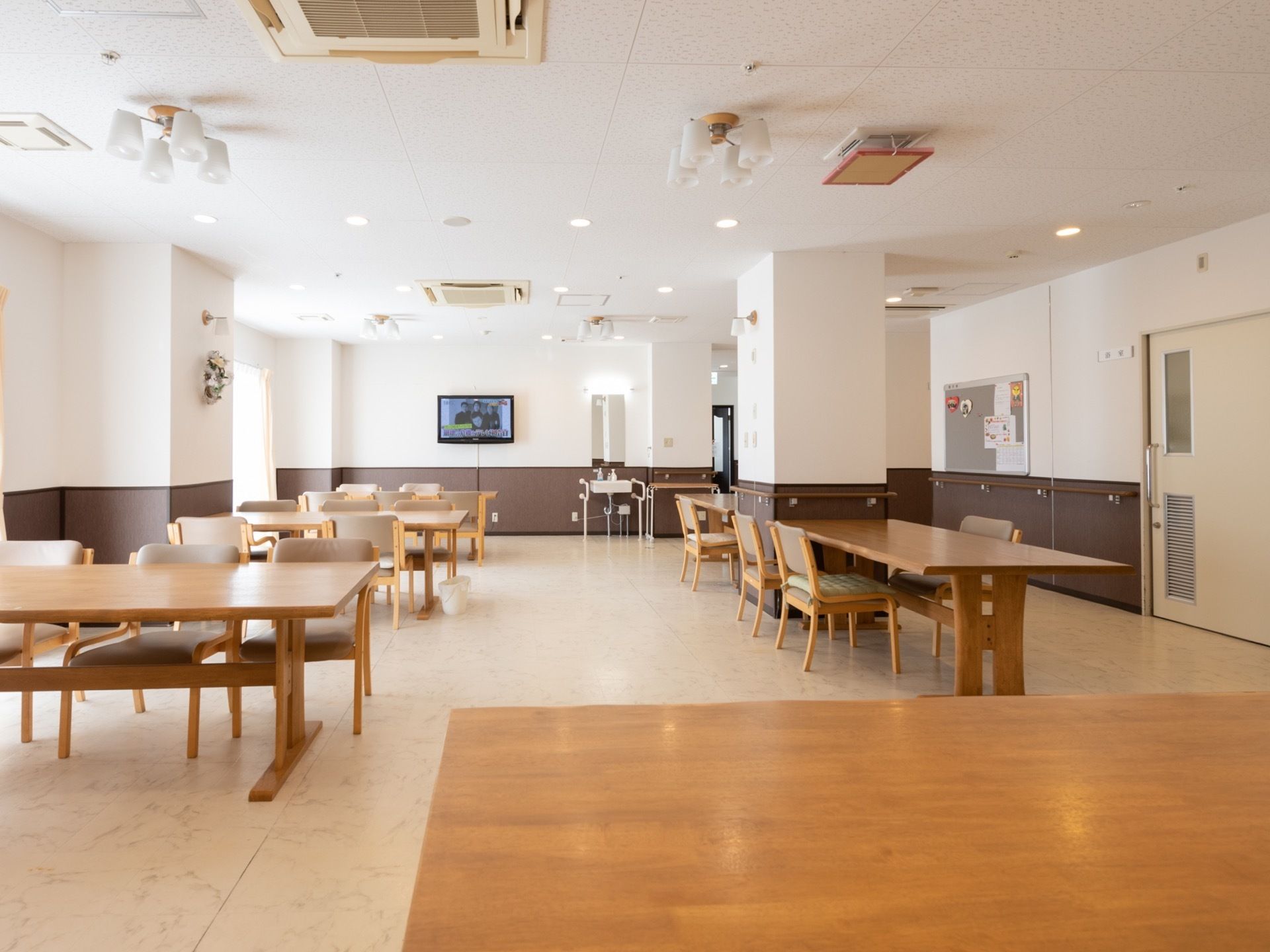 明るく広い食堂に､木製の大きめのテーブルセットが並んでいる。正面にはテレビがあり、フットレスの洗面台も見える。