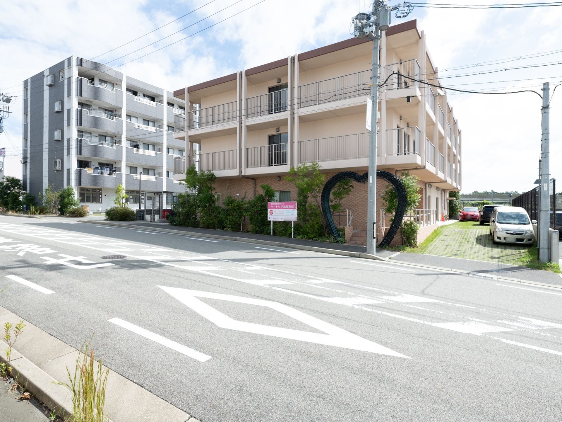 建物は落ち着いたピンク色の鉄筋3階建てで、テラスには白いフェンスがめぐらされている。緑の植木の横の階段にハート型のアーチがある。