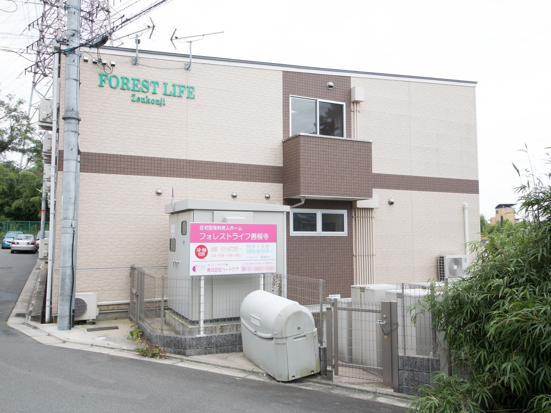 歩道と施設の間はフェンスで区切られている。フェンス内に施設の案内看板が建てられている。奥に山麓がみえる。