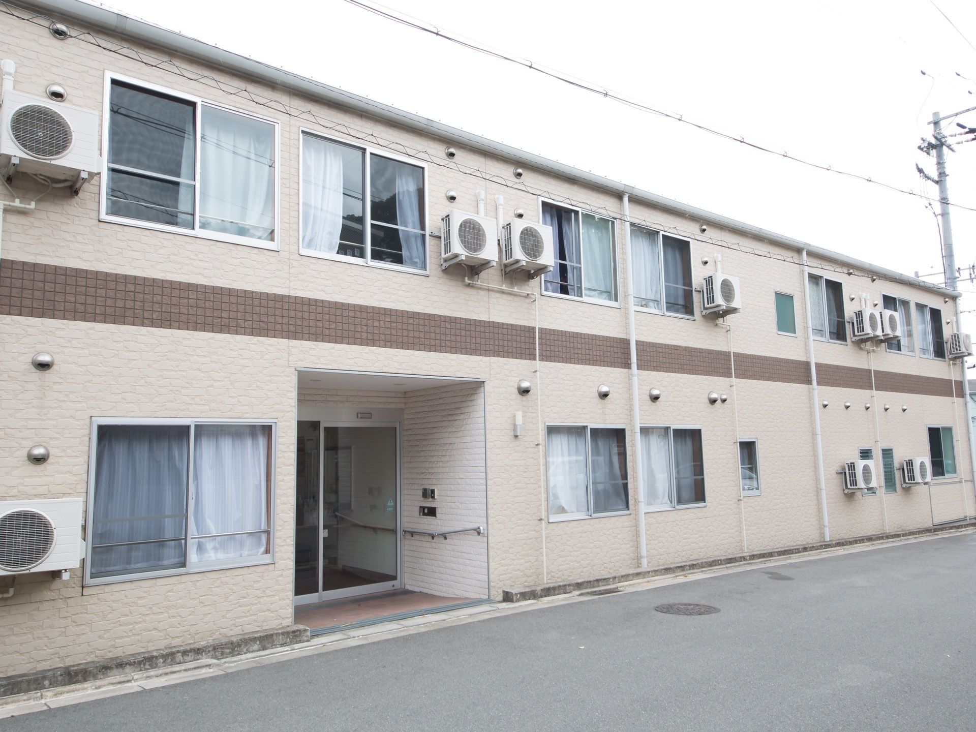 白を基調とした建物で、中心に茶色のラインが入っている。玄関横の外壁にはインターホンが設置されている。