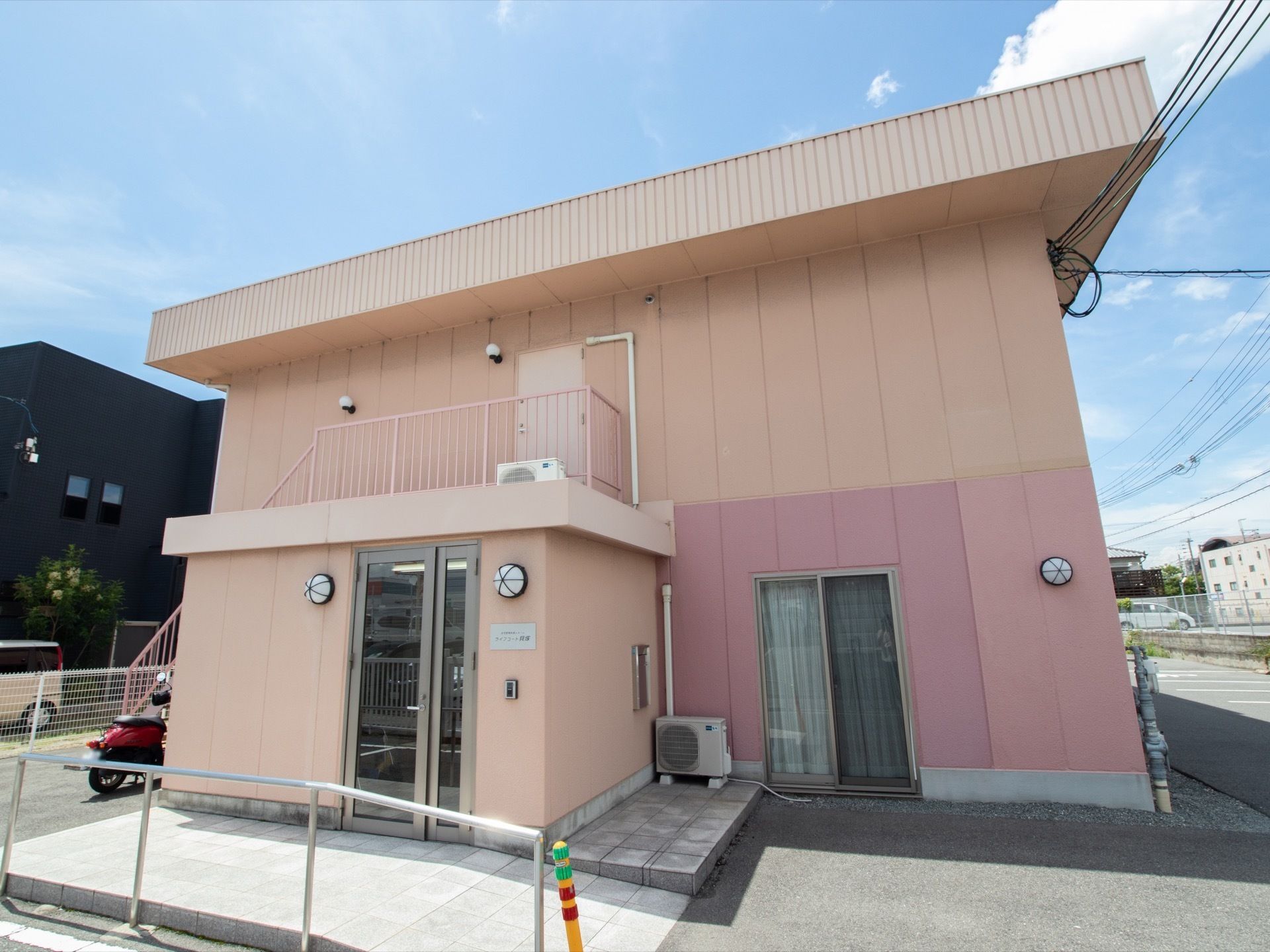 ピンク色の外壁の建物で、地上２階建てとなっている。駐車場から入口に手すりつきのスロープがつながっている。