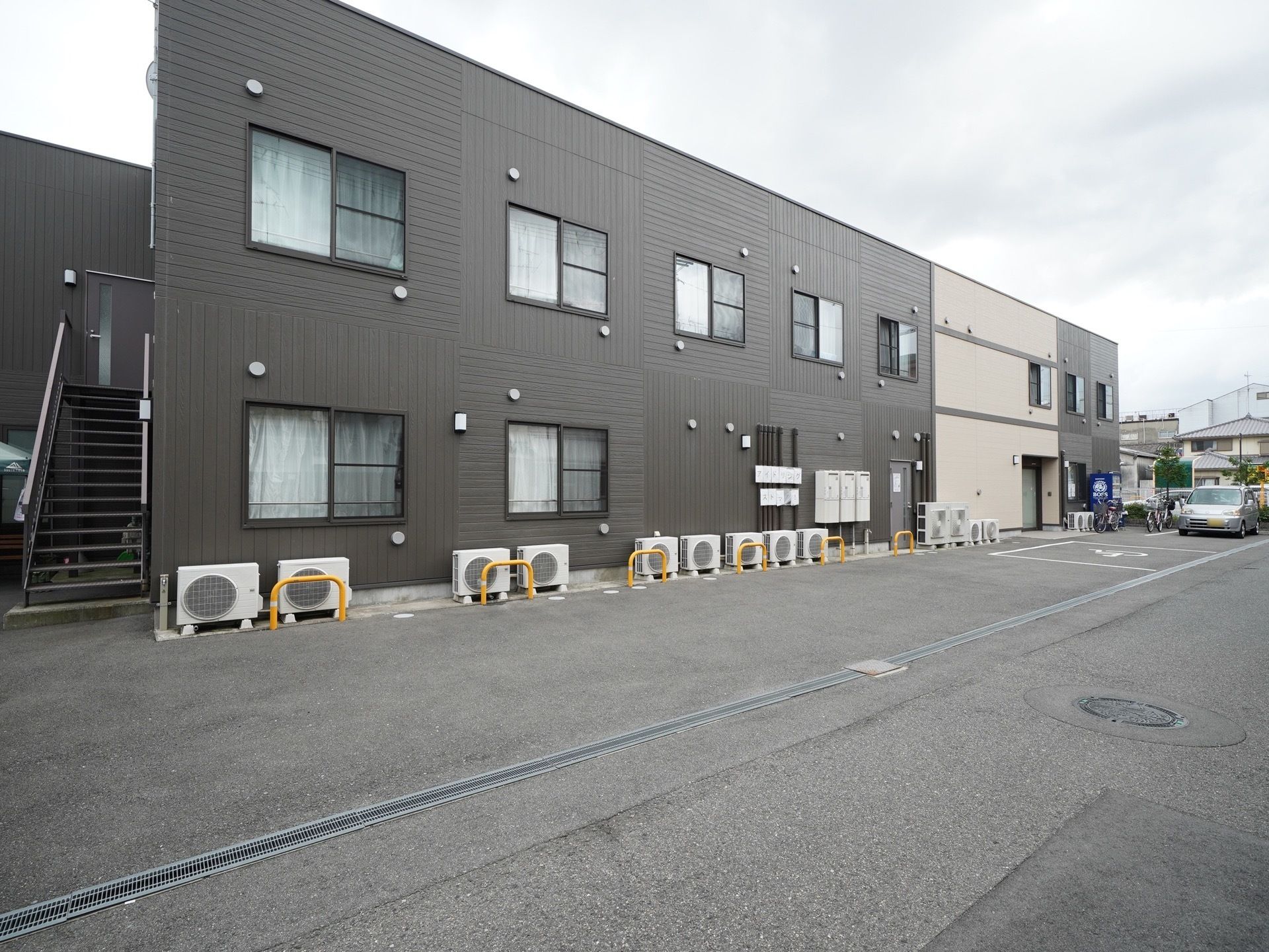 2階建ての建物になっており、シックなカラーの外観になっている。外にはエアコンの室外機が置かれている。