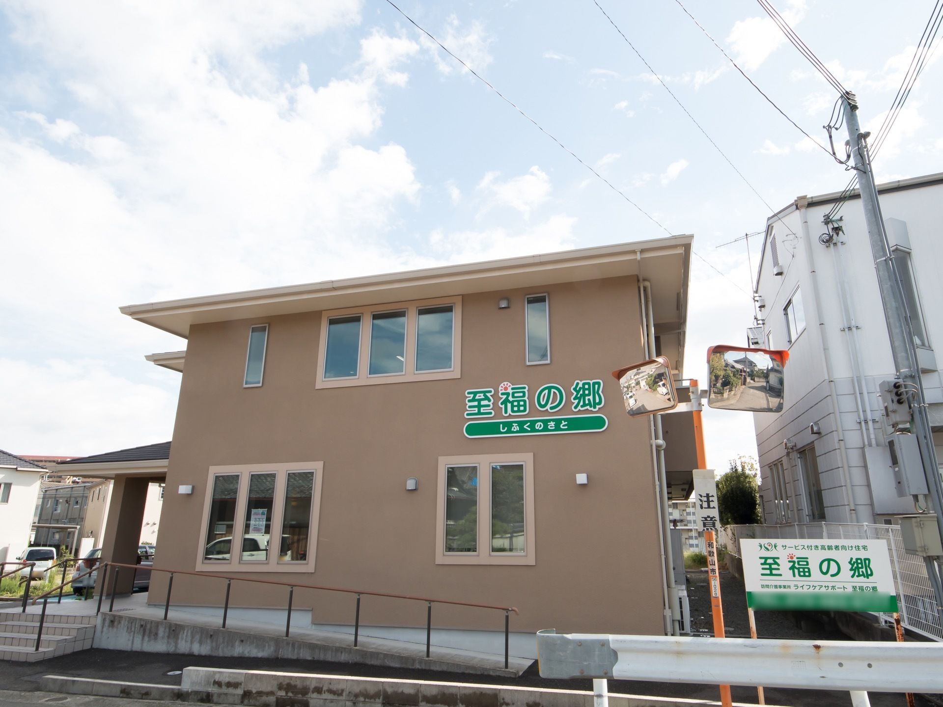 施設は茶色い外壁で２階建てである。施設周りの一部は白いガードレールがあり、その近くには階段とスロープが設置されている。