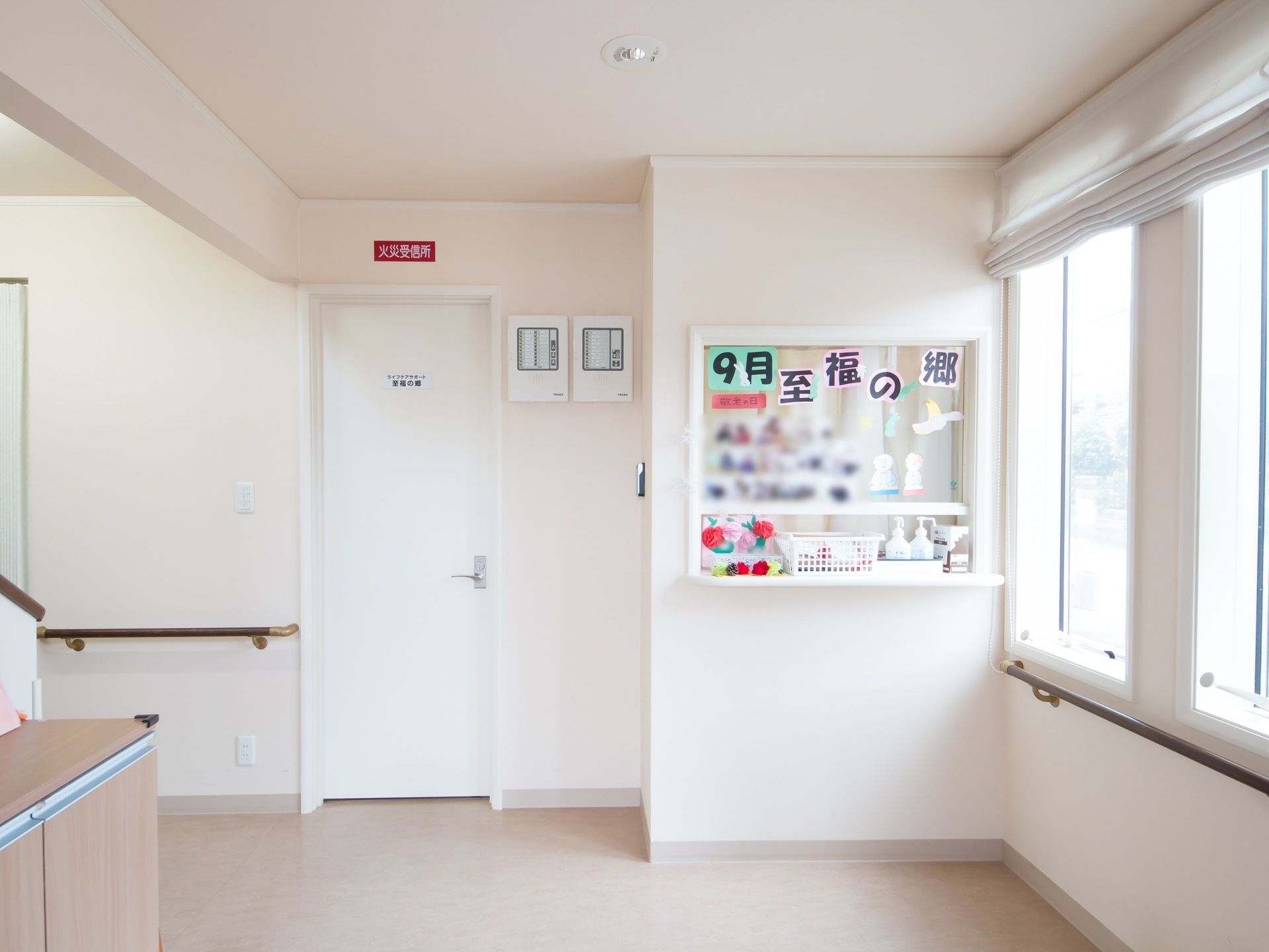 通路の壁は白く、床は薄いベージュである。窓には白いブラインドカーテンあり、その下には手すりが設置されている。