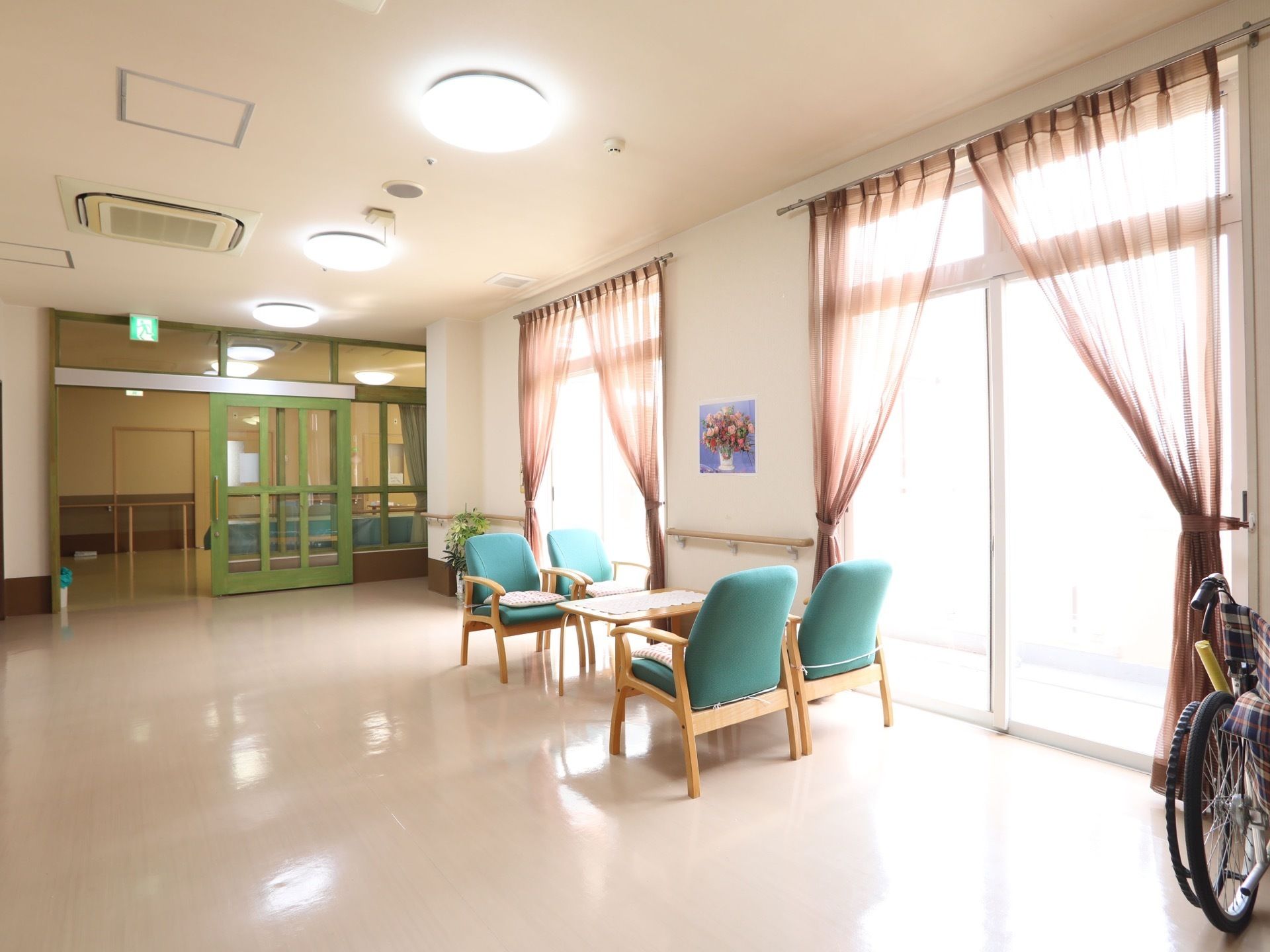 薄いピンクのカーテンが掛けられた窓が柱を挟んで２つ設置されている。窓の間の柱には花の絵が飾られている。