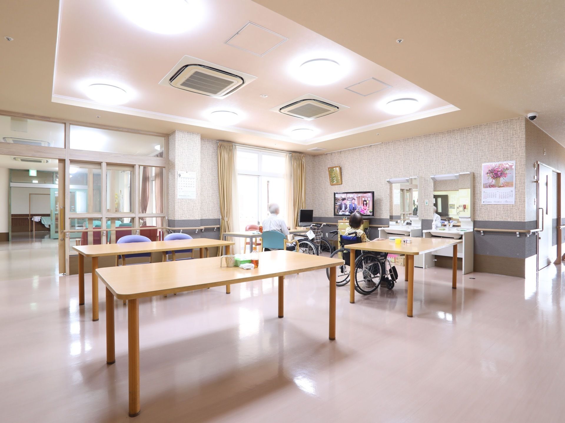 天井の照明が床に反射して光っている。腰高の細長い木製テーブルが３台ほど並んでいる。奥にはテレビがある。
