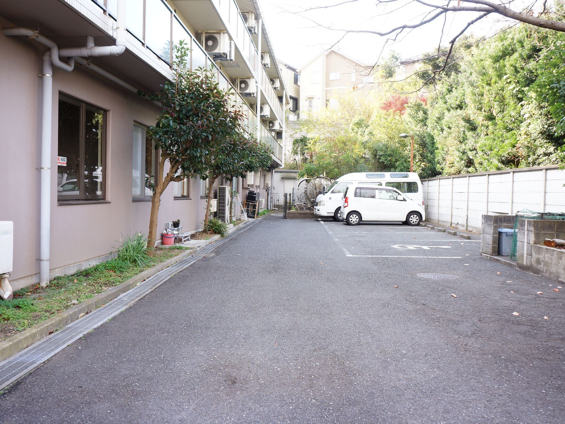 建物の隣に駐車場がある。車椅子専用のスペースもある。脇にはゴミ捨て場がある。駐車場の後ろには塀があり、大きな木々がある。