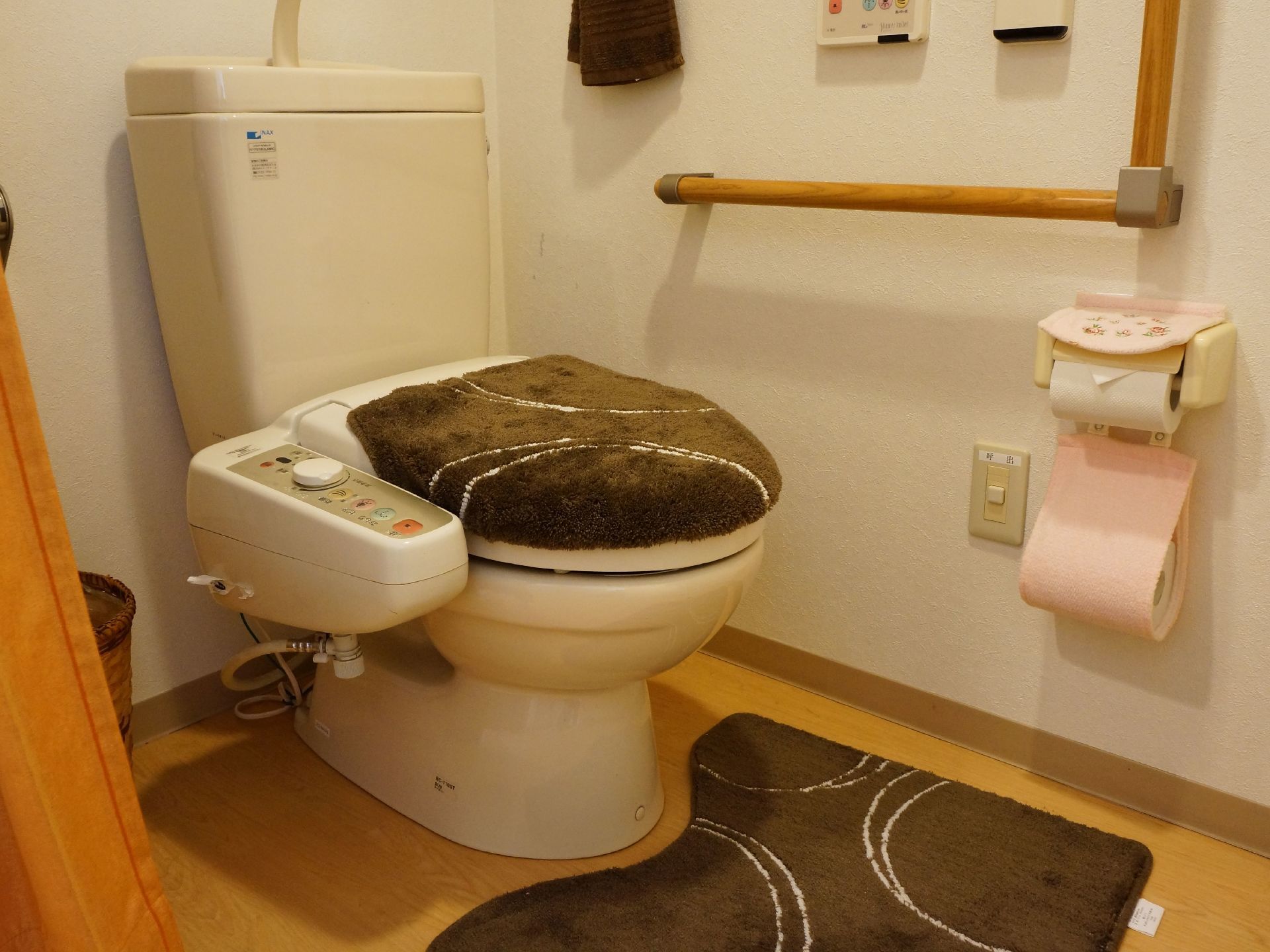 茶系に円が描かれたお揃いの便座カバーとマットがある。壁には手すりが設置され、温水洗浄機能のボタンがある。