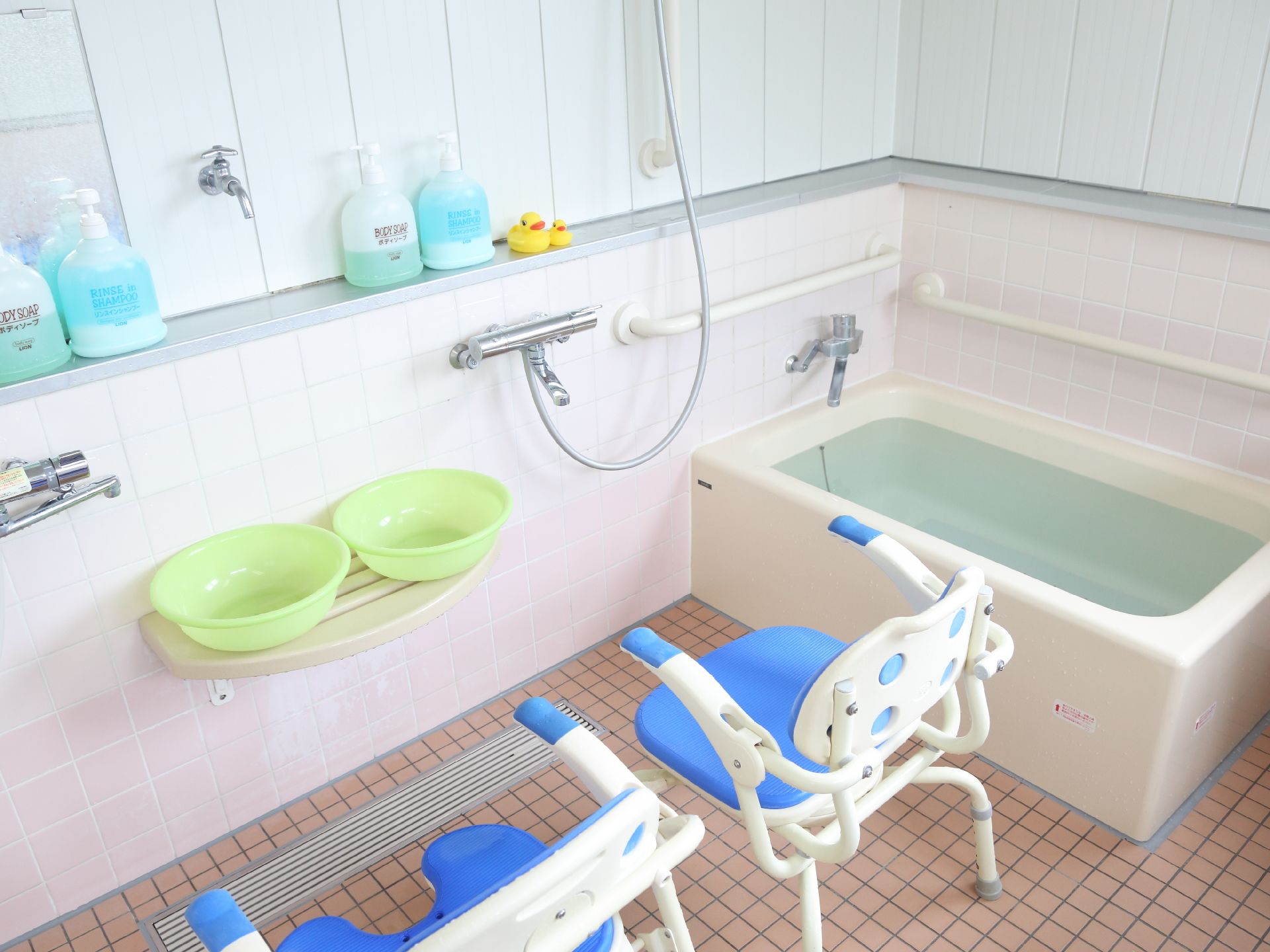 浴室の一角の写真。浴槽の隣にはシャワーが設置され、背あてとひじ掛けの付いた椅子が置かれている。周りの壁には手すりが付いている。
