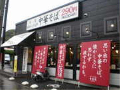 ラーメンチェーン店の建物の外観である。建物の壁の色は茶色系で、入口には屋根が付いている。周囲には看板が並べられている。