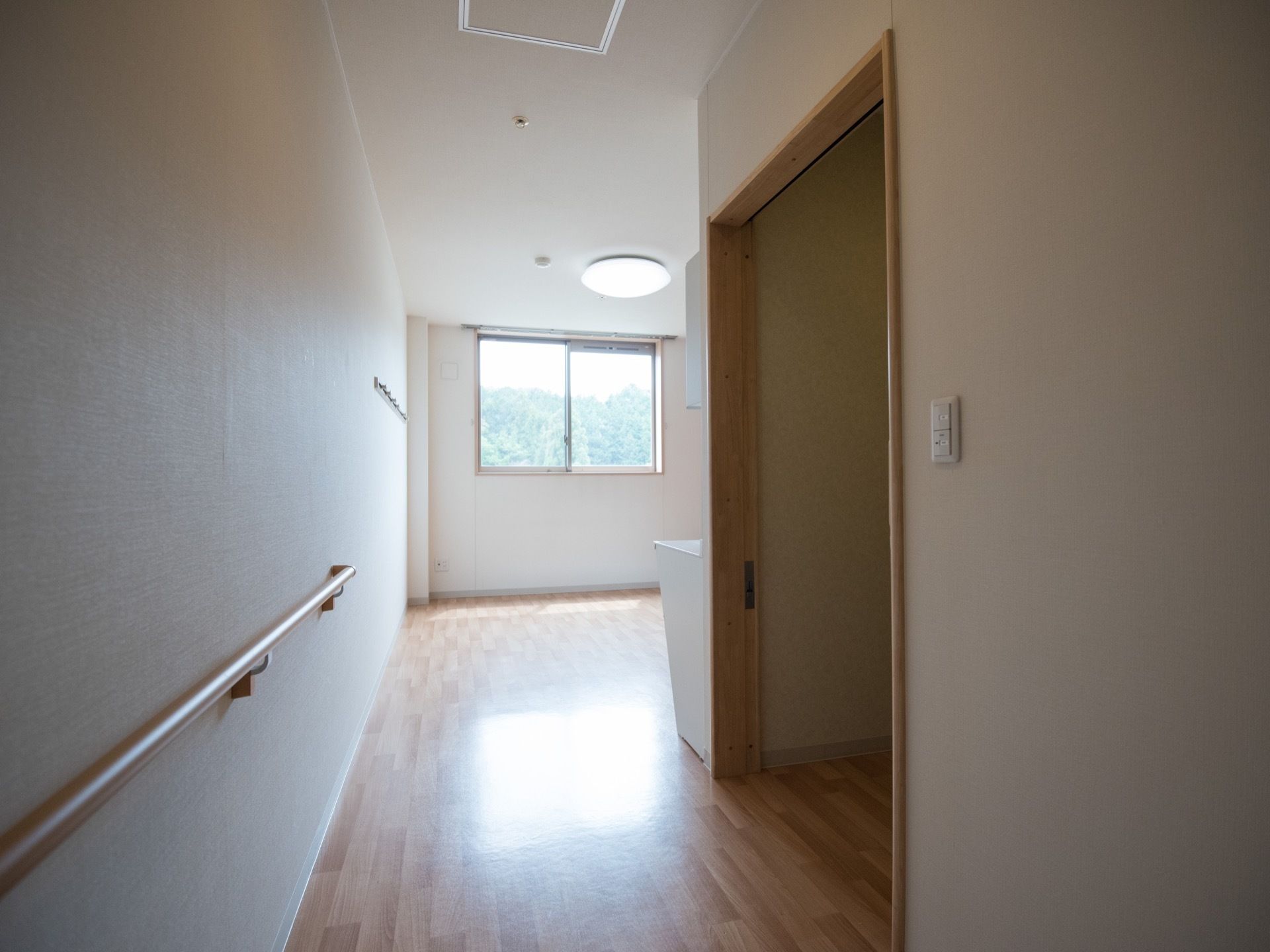 奥行きのあるフローリングの部屋がある。入口近くの壁には手すりが設置されていて、突き当たりには窓がある。