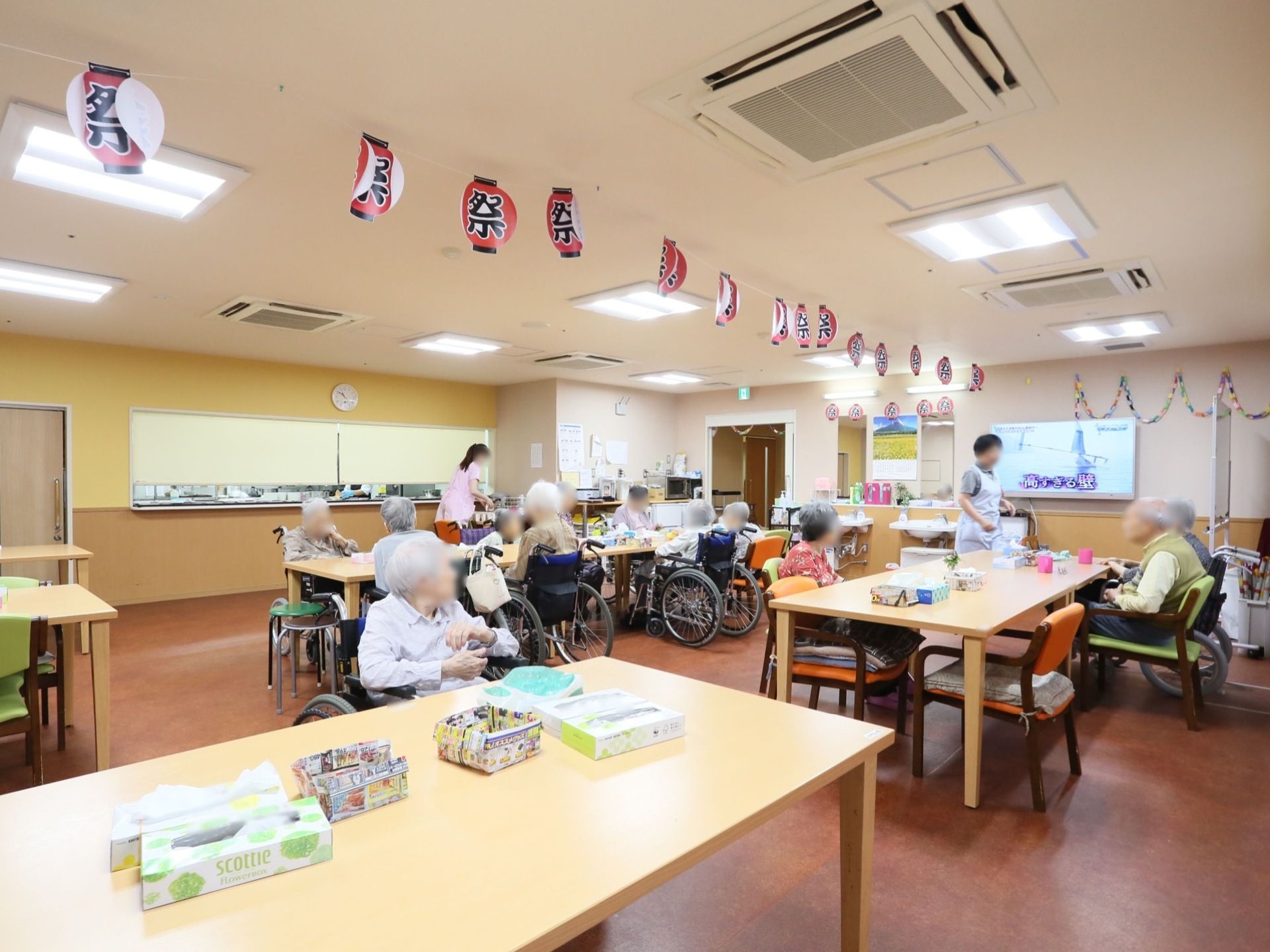 共有スペースでは、男女の入居者様や車椅子をご利用の方がくつろがれている。介助する2名の女性スタッフの姿が見える。