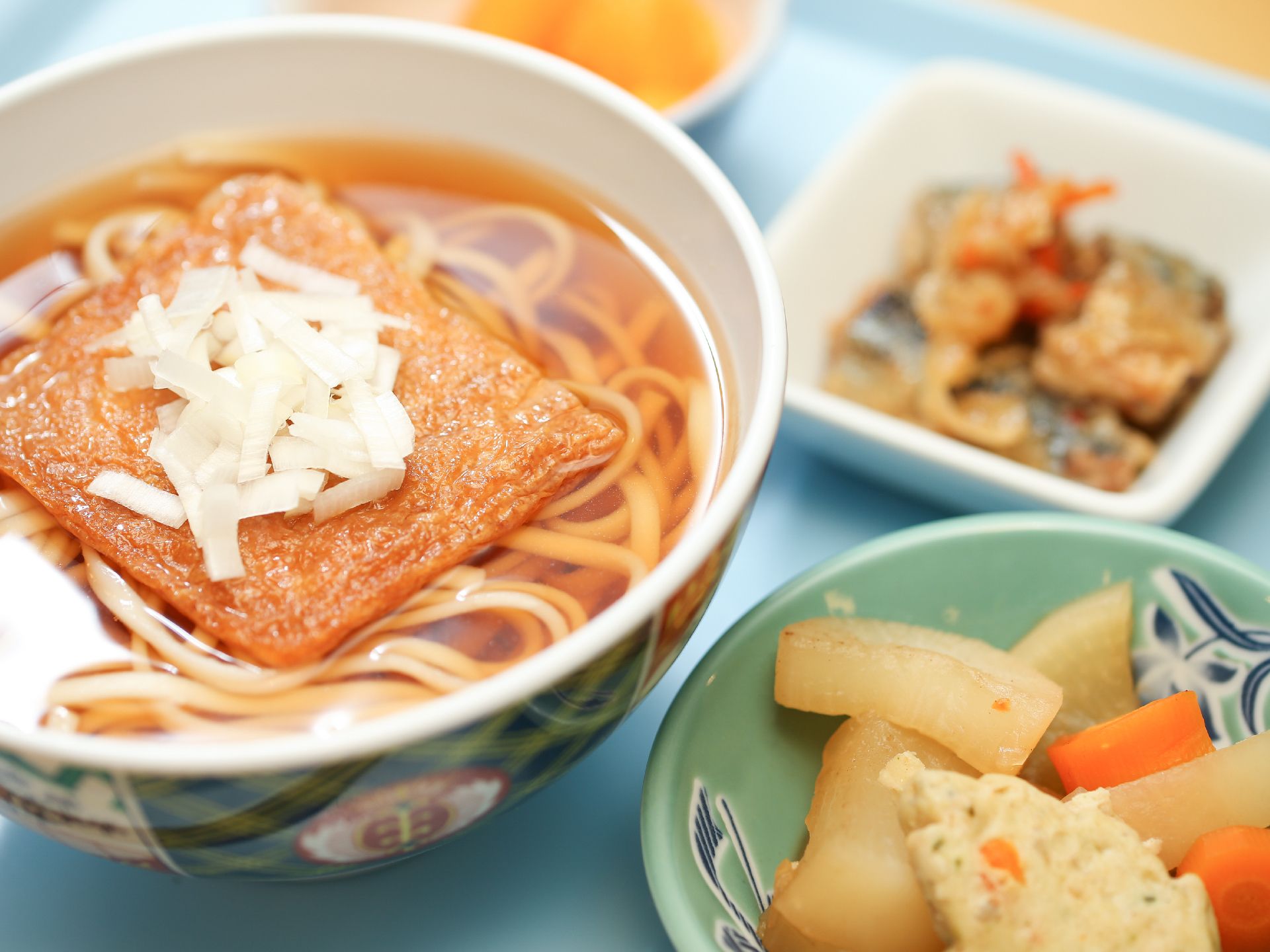 介護施設の食事は栄養バランスを考えている。きつねうどん、大根の煮物、魚の南蛮漬け、フルーツが並べられている。