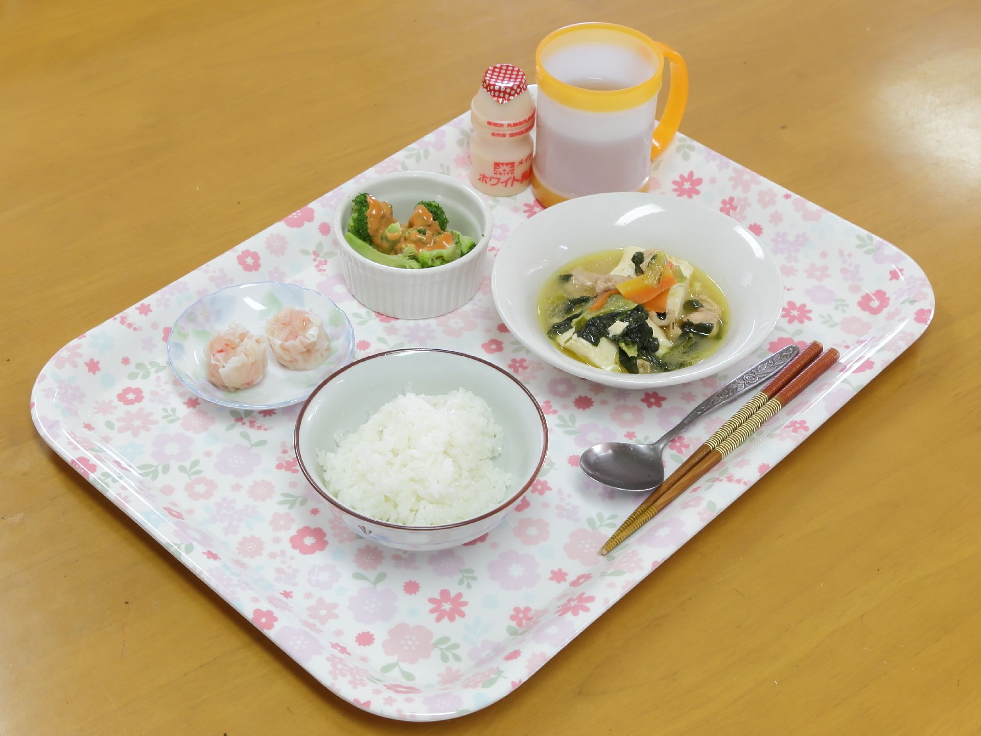 花柄のトレイにご飯や具が多めの汁物、おかずが並べられている。乳酸飲料やお茶の入ったマグカップも置かれている。