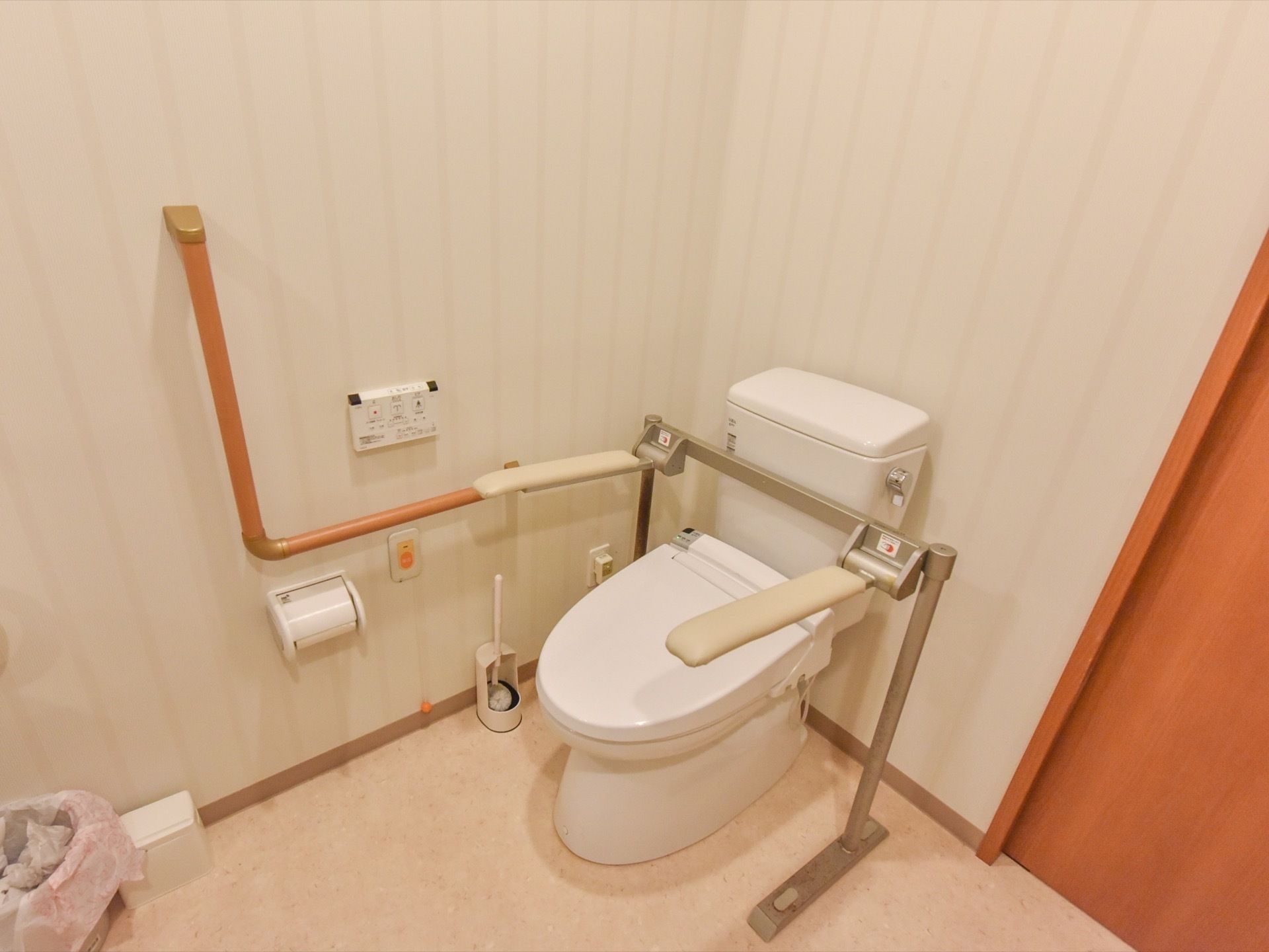 壁に茶色の手すりが付いている。便器の両脇には跳ね上げ式の手すりが備わっている。オレンジ色のボタンは緊急呼び出しボタンである。