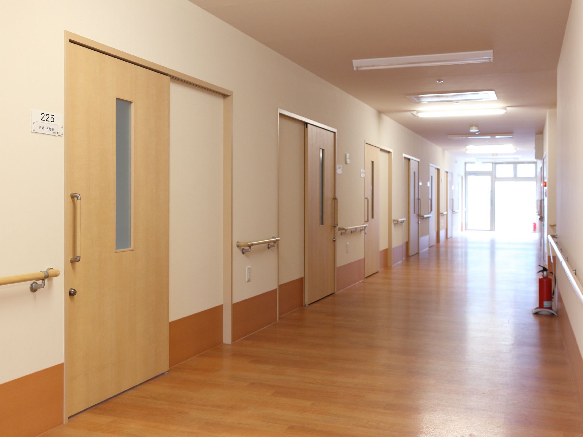 廊下に居室が並んでいる。居室には部屋番号と名前が表示されている。扉は横開きで、壁に手すりがついている。