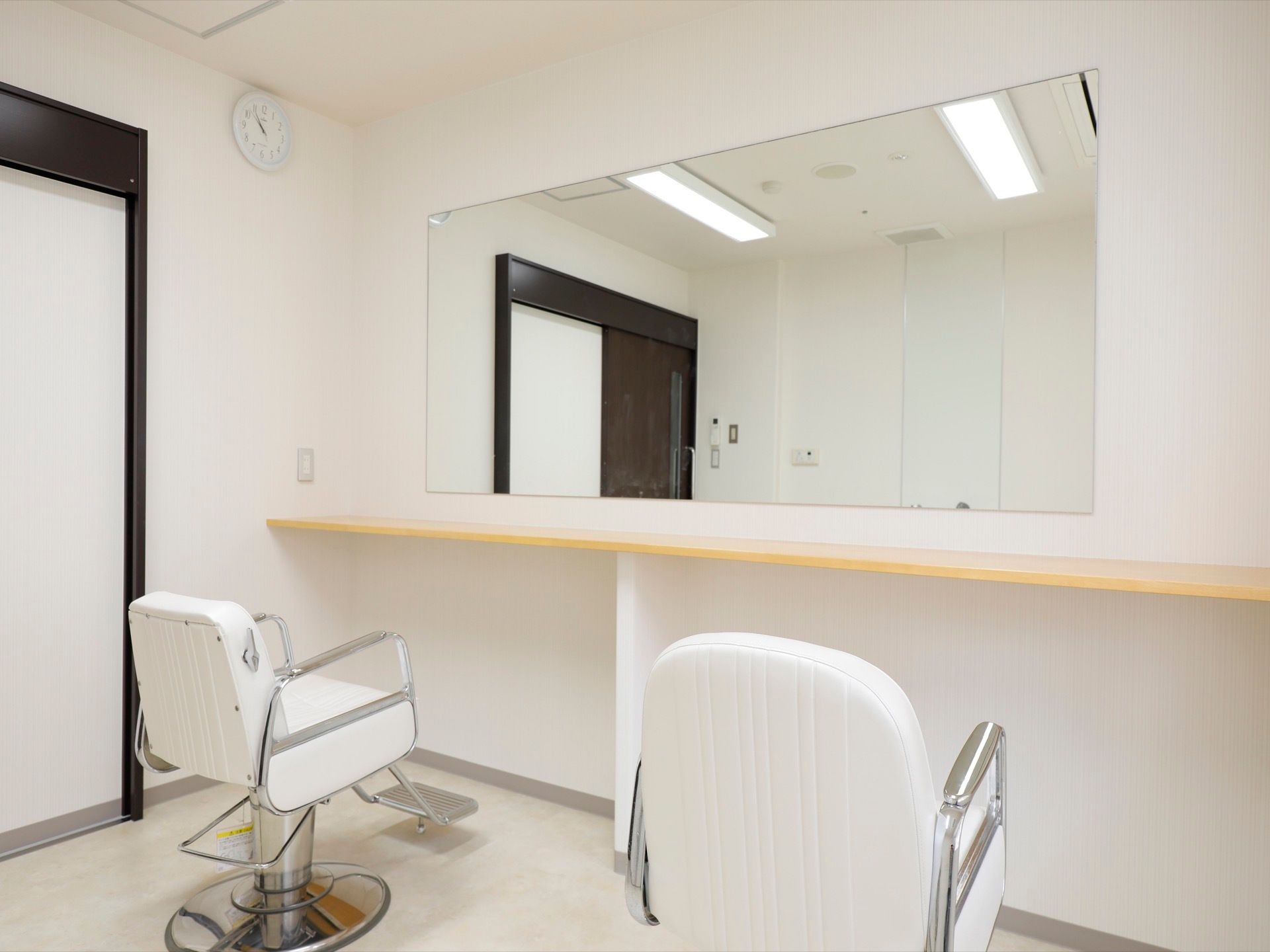 白を基調とした清潔感のあるヘアーサロンに、椅子が２脚置かれている。大きな鏡があり、壁には時計がかかっている。