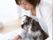 日当たりのよい明るい室内でくつろぐ白いシャツを着た女性。腕の間にはグレーと白の毛色の小型犬が座っている。