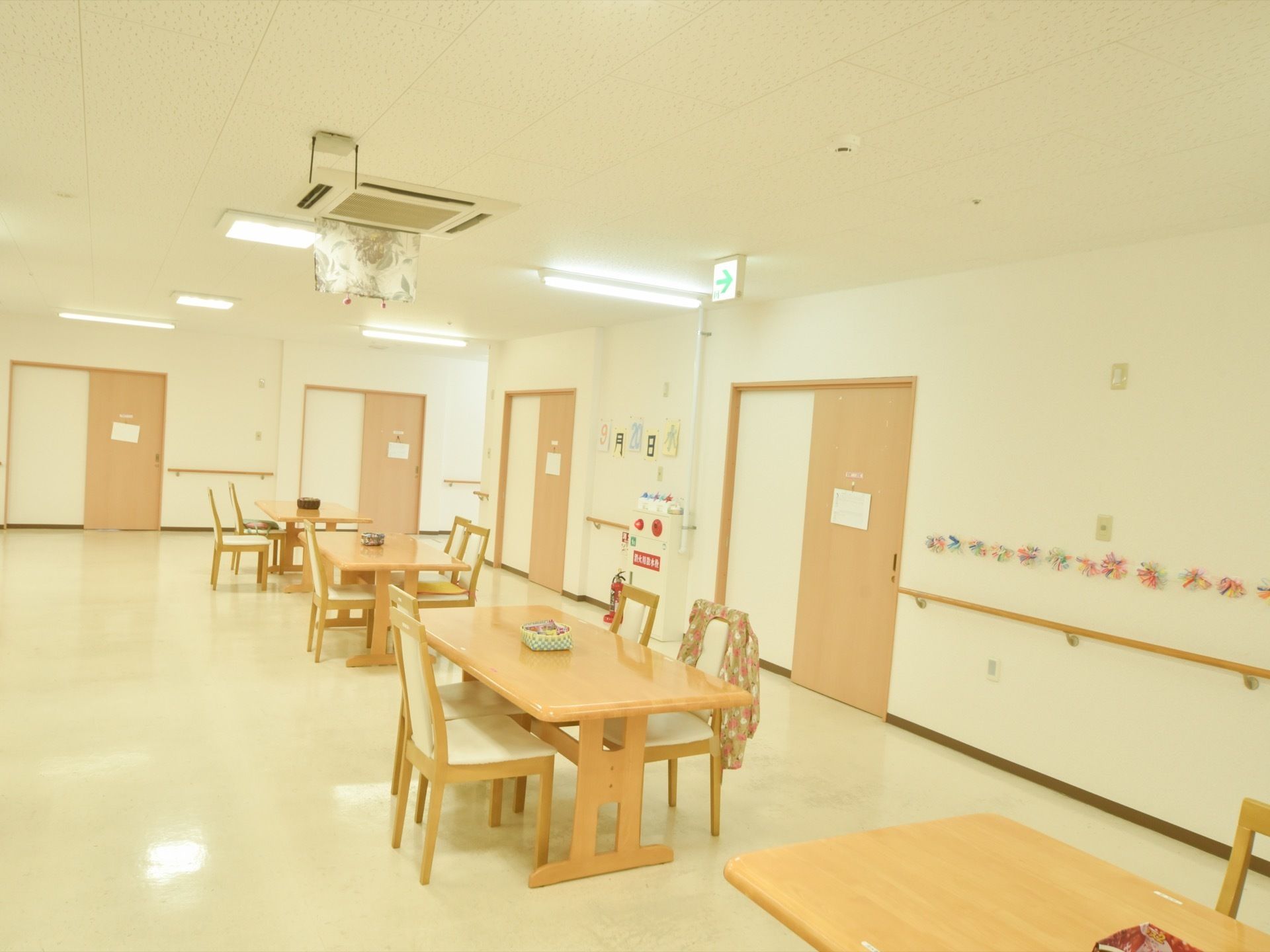 クリームとライトブラウンが基調の広々とした共有スペースの様子。テーブルセットが間隔を保って置かれており、壁には手すりが付いている。