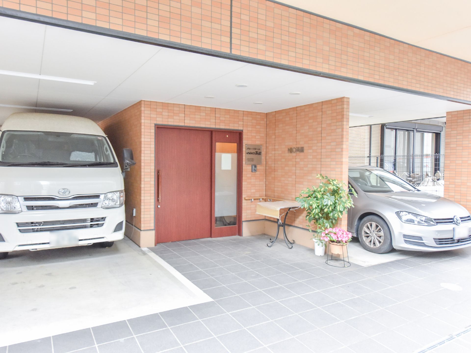 施設のエントランスは、1階の駐車スペース一角にある。入口の壁にも手すりが設置されている。また、花や植物が飾られている。