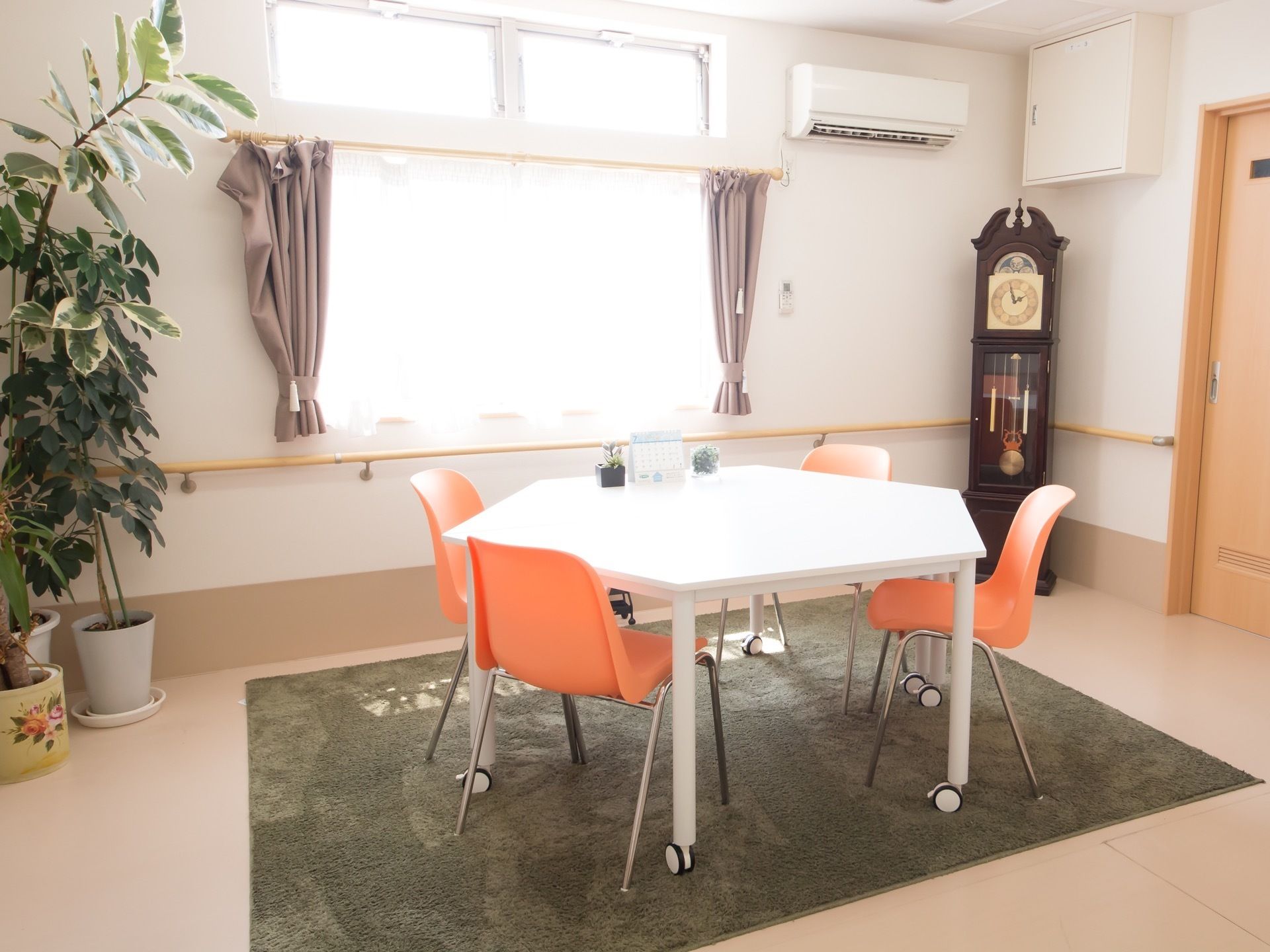 日差しの差し込む明るい部屋である。白い五画形のテーブルにオレンジ色の椅子がセットされている。奥に大きな時計が置かれている。
