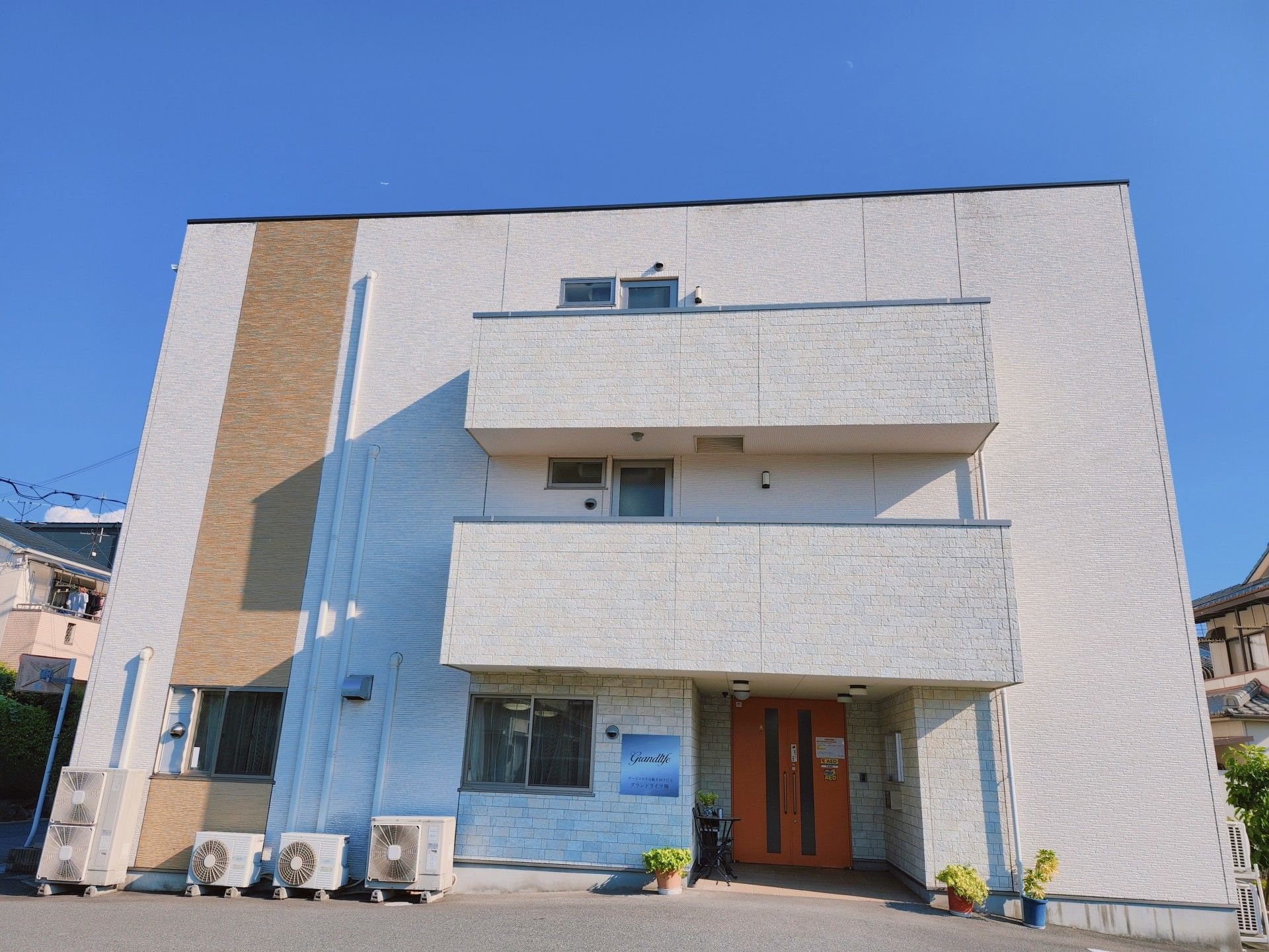 白色をベースに、縦にベージュのラインが入った３階建ての建物がある。施設入口の前には駐車スペースがある。