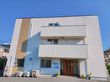 白色をベースに、縦にベージュのラインが入った３階建ての建物がある。施設入口の前には駐車スペースがある。