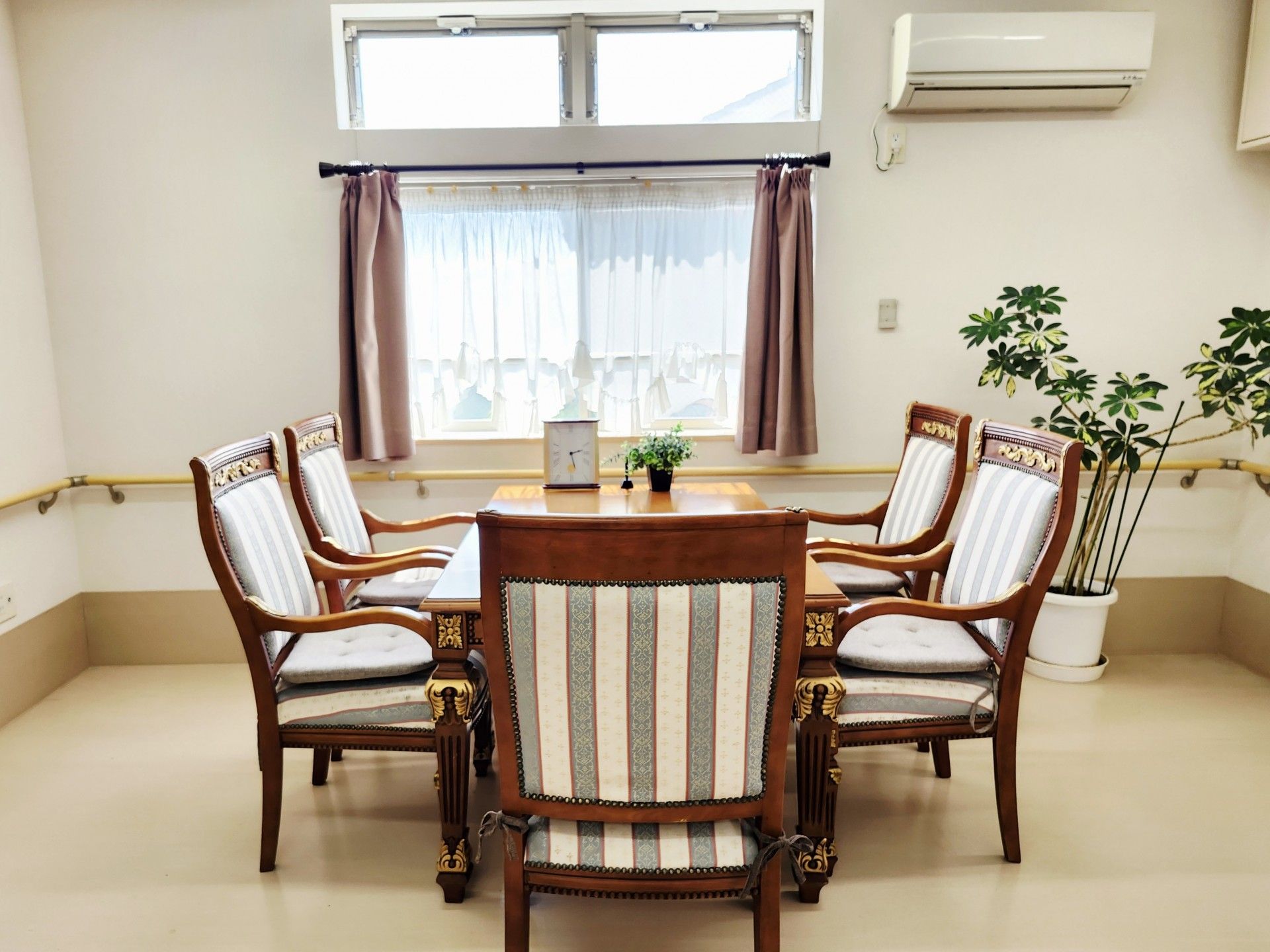 日差しの差し込む明るい部屋である。白い五画形のテーブルにオレンジ色の椅子がセットされている。奥に大きな時計が置かれている。