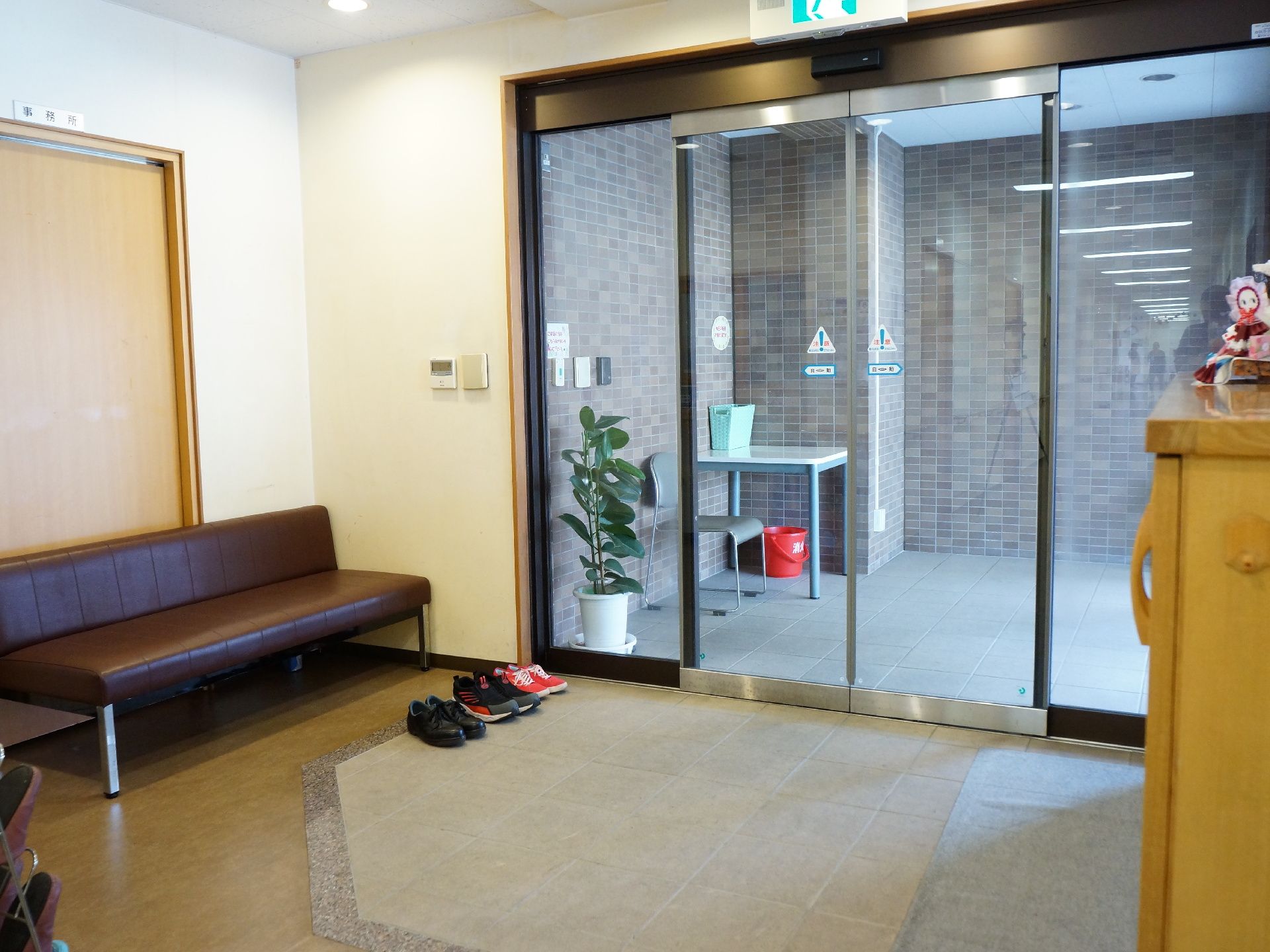 玄関の扉は自動ドアになっている。たたき部分に靴が置かれている。玄関には事務室が隣接していて、扉の前にベンチが設置されている。