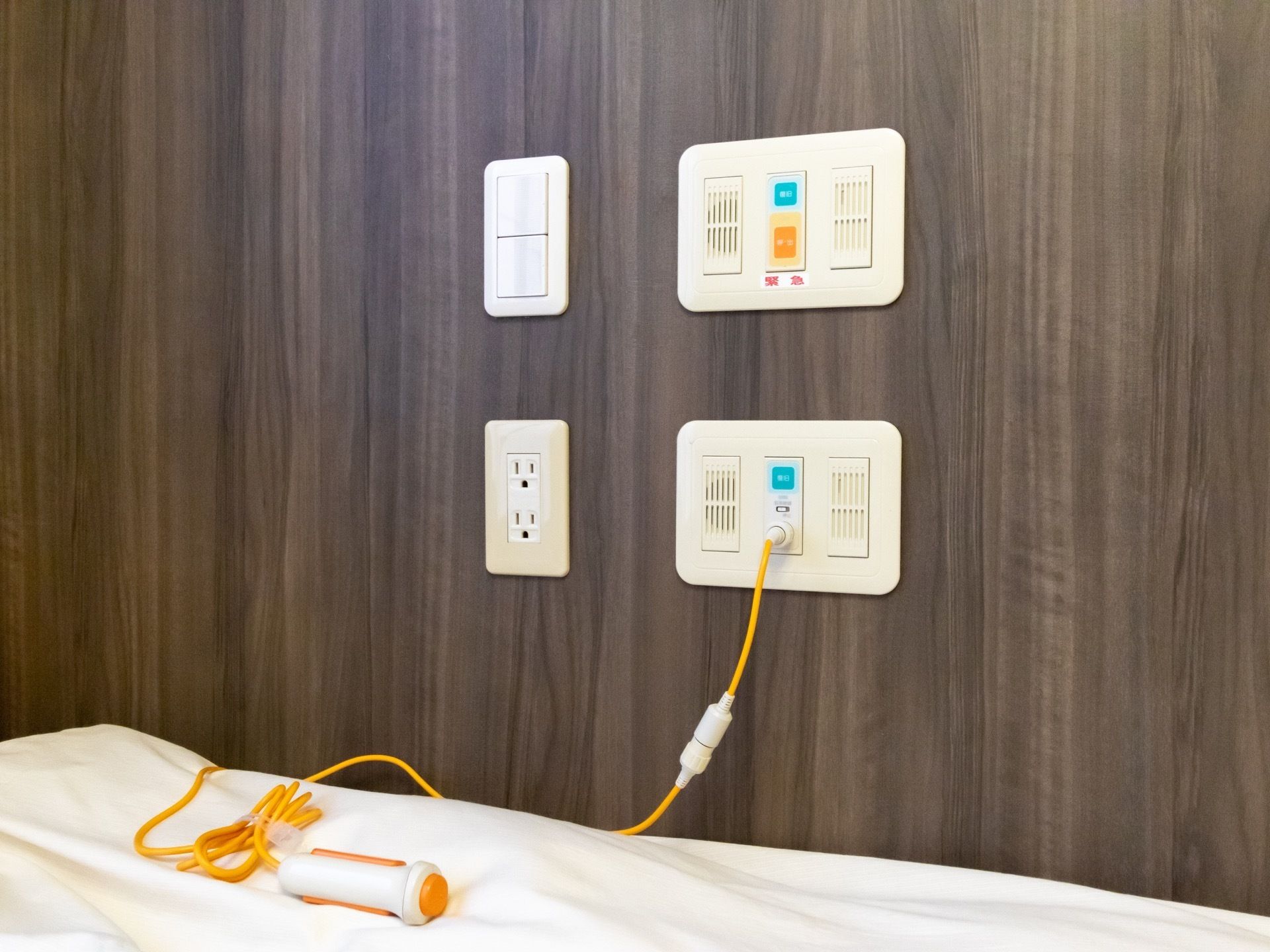 グレーの木目調の壁にナースコールがある。側にはコンセントや、電気のスイッチなども設備されている。