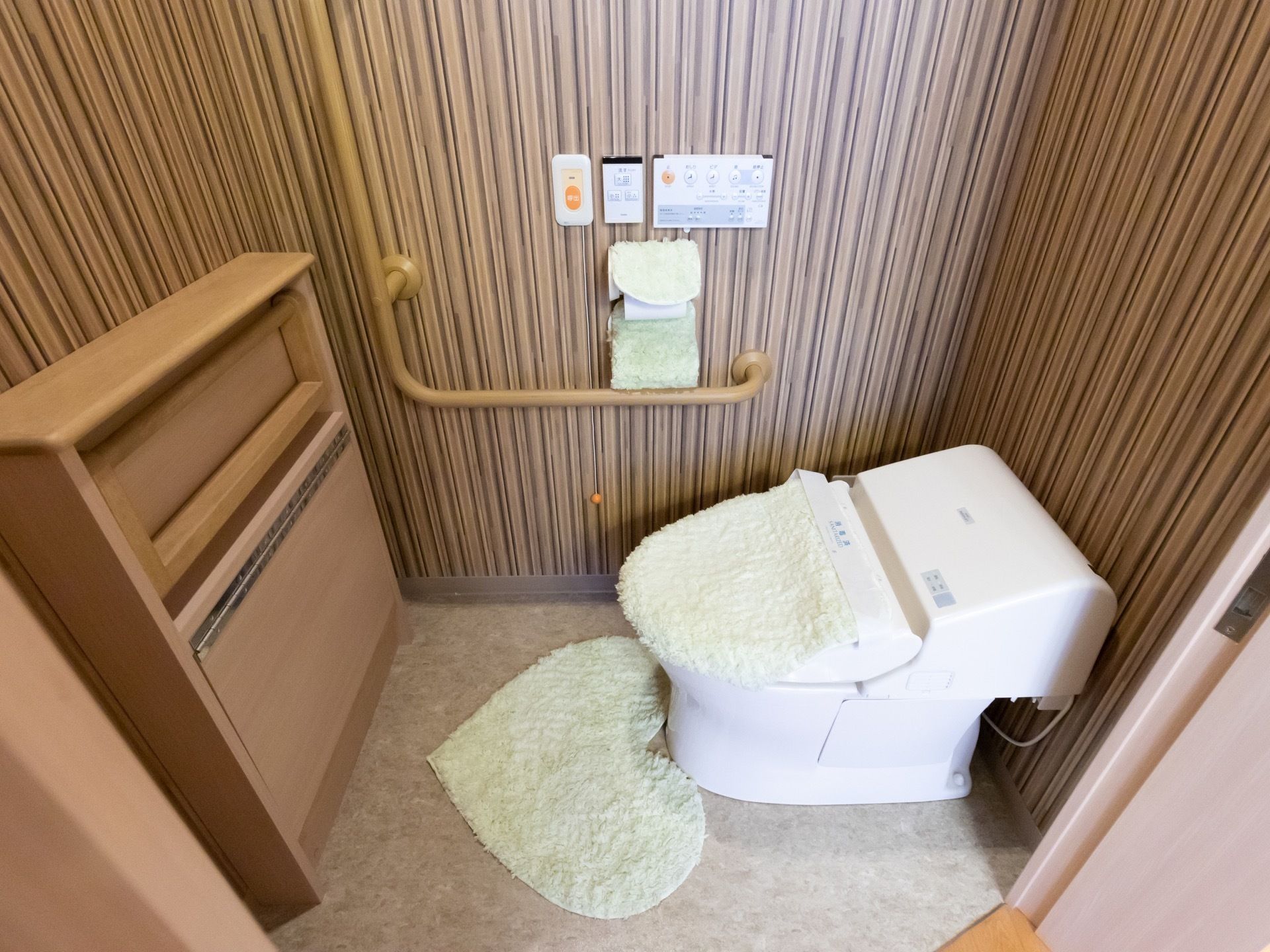 トイレには温水洗浄便座が設置されており、床にはトイレマットがある。壁にはL型の手すりや呼び出しボタンなどが備えられている。