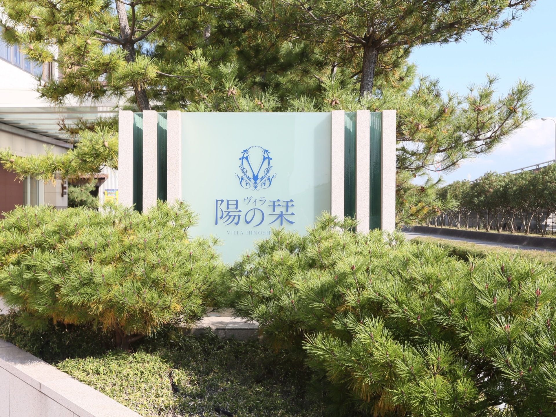 薄いブルーの看板が立っており、施設名が書かれている。周りには木が植えられていて、近くには道路がある。