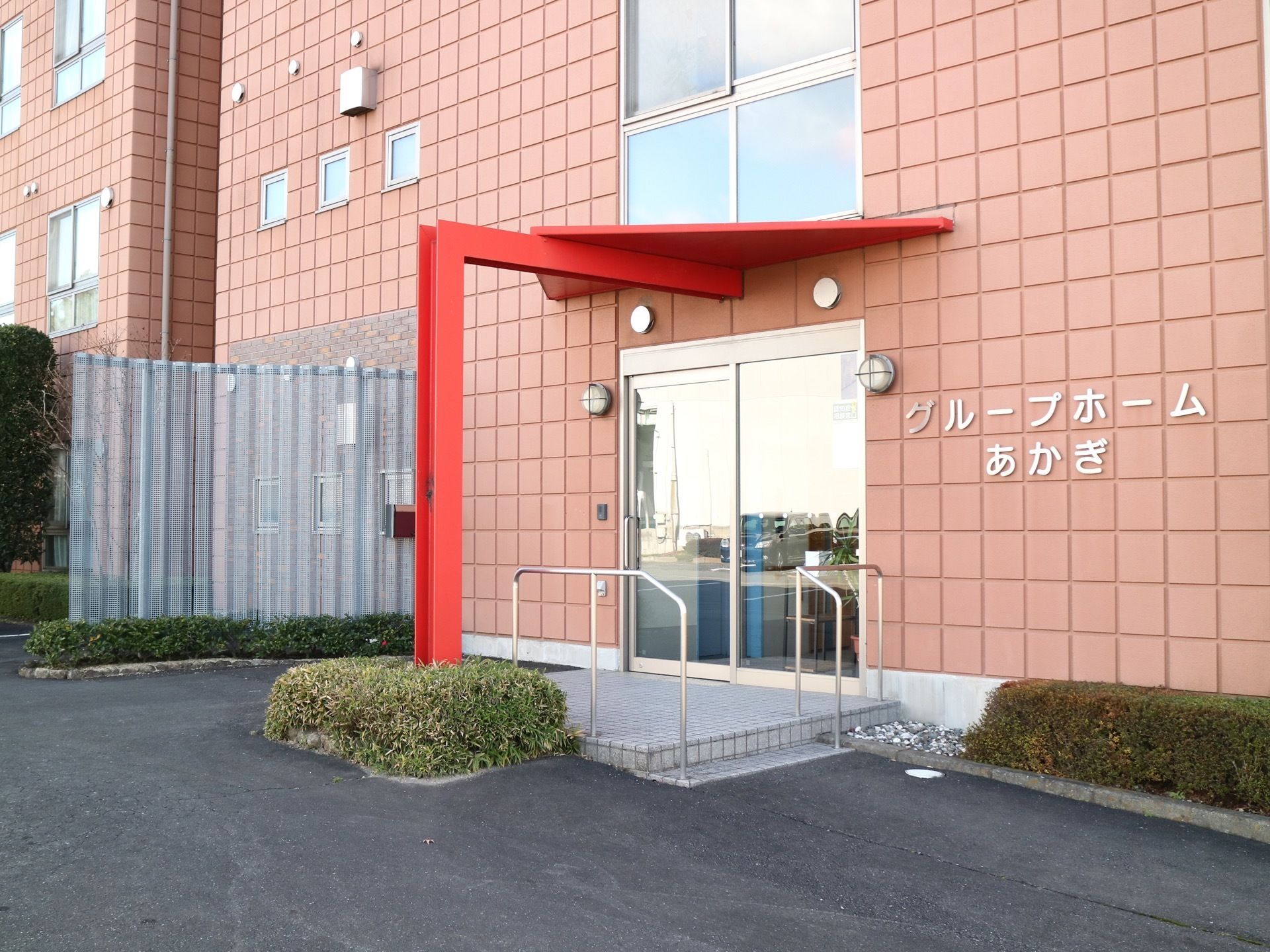スライド式のドアの玄関で、右側の外壁に白い文字で施設名が書かれている。左側の外壁にインターフォンがある。