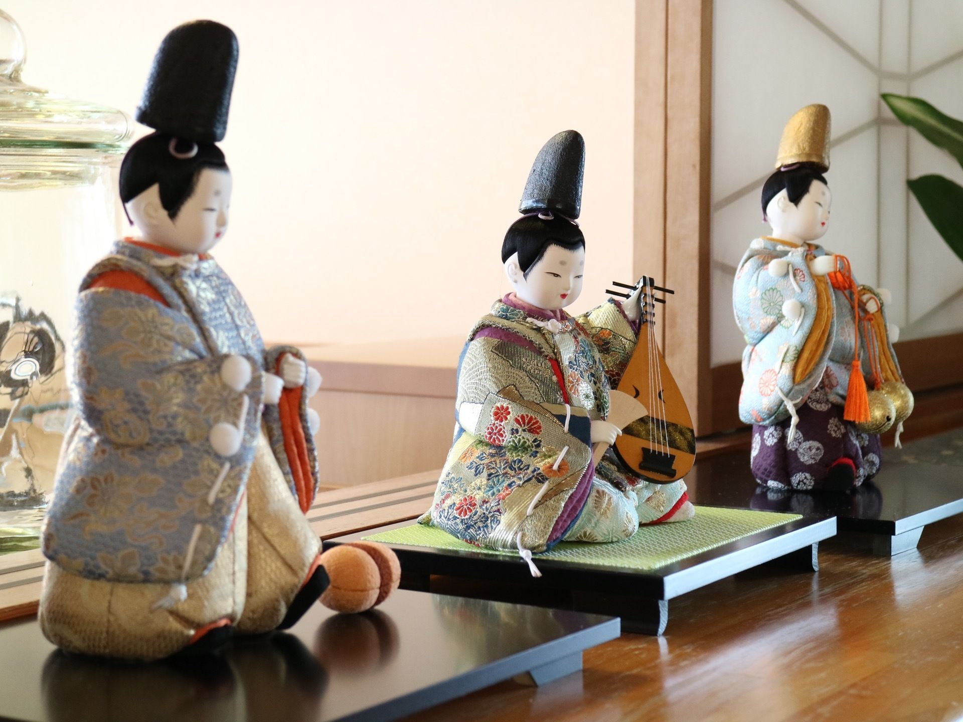 茶色の棚の上に和人形が３体並べて飾られている。その右側には、人形と同じ位の高さの観葉植物が置かれている。