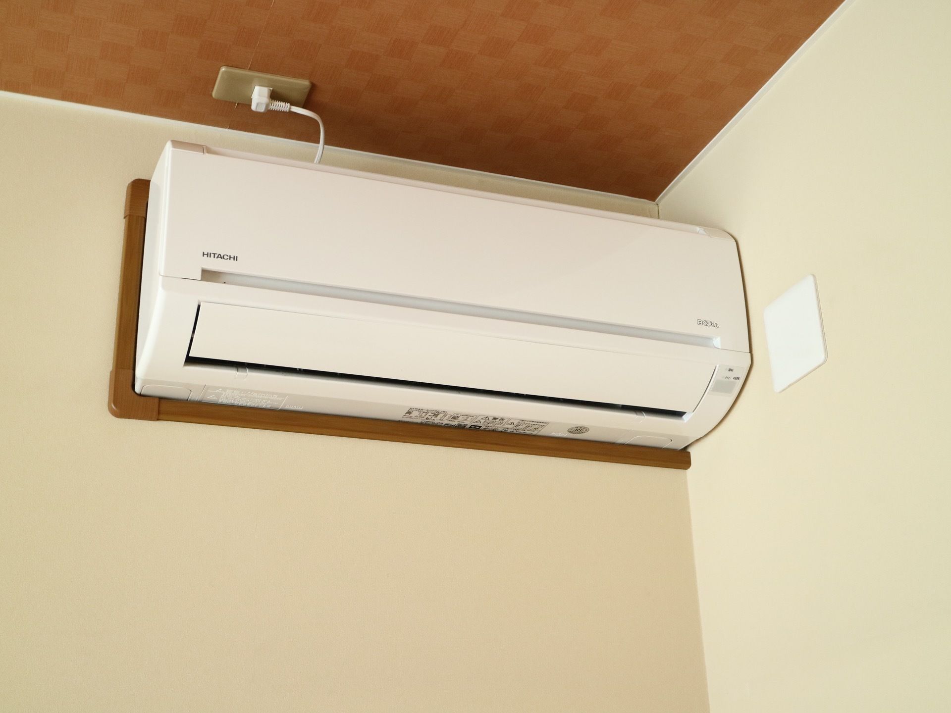 白い壁の隅にエアコンが１台取り付けられている。茶色い天井にコンセントタップがあり、電源が繋がっている。