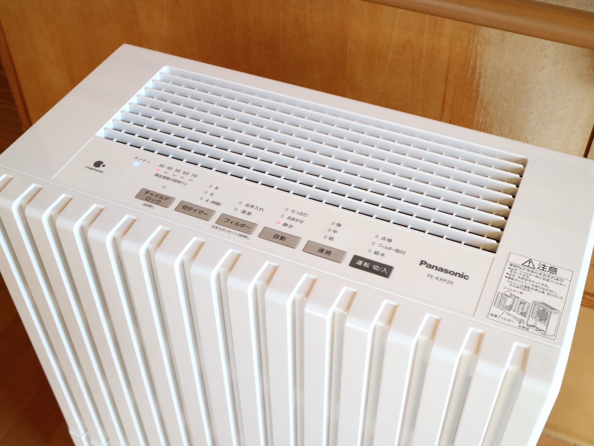 茶色い壁の前に白い加湿器が１台置かれている。稼働中のようで、上部にある表示面のランプが点灯している。