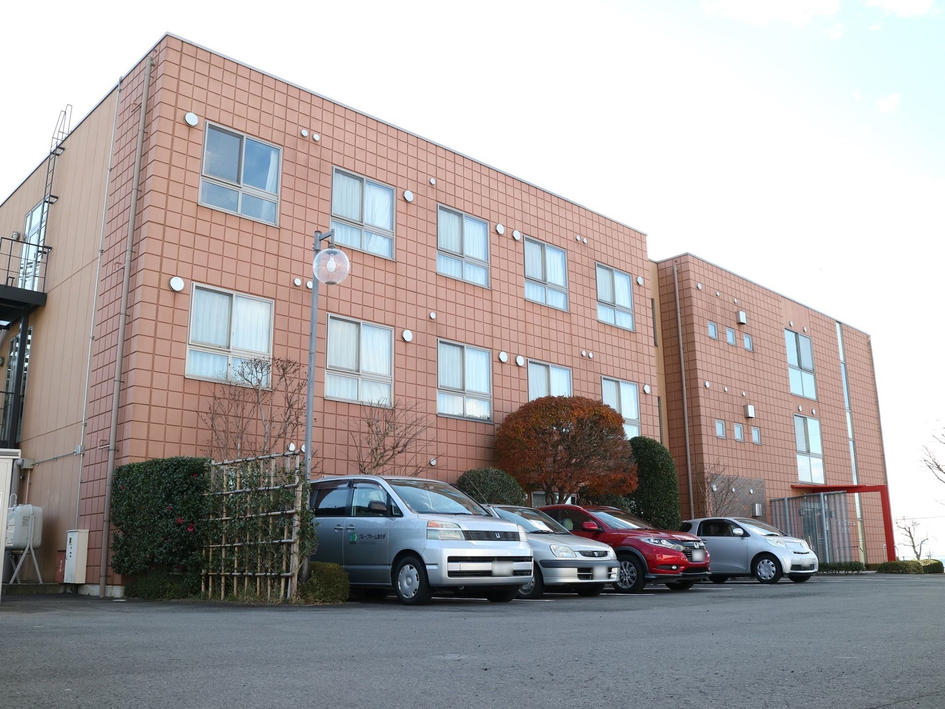 ３階建ての施設で、建物に沿うようにさまざまな高さの樹木が植えられている。その前は駐車場になっており、車が停まっている。