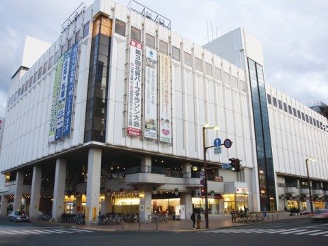通りに面した大型商業施設。建物にはイベントの垂れ幕が掲げられている。人や車の通りも多く、賑やかな場所である。