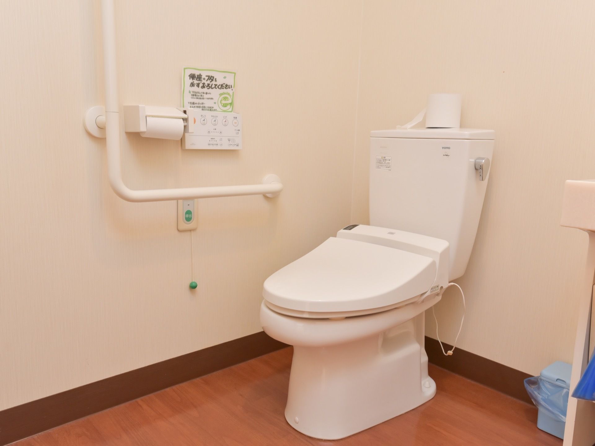 木目調のフローリングの床のトイレ。横に小さなごみ箱が置かれており、壁には紐付きの呼び出しボタンや張り紙がある。