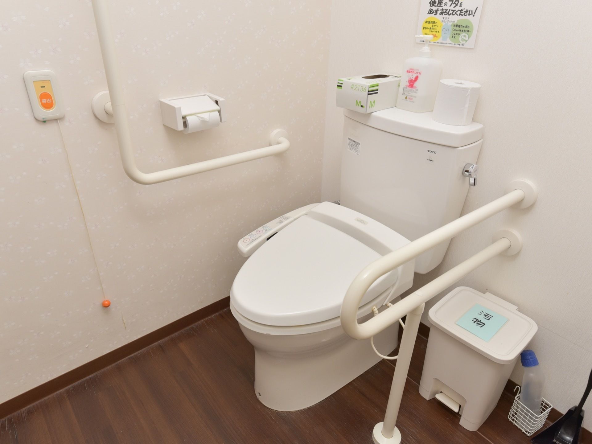 壁や便器の脇に、手すりが設置されている。ナースコールが備わっている。消毒液や汚物入れも置かれている。