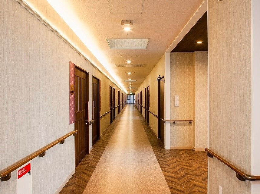 廊下はまっすぐに長く続き、両側に居室のドアが並んでいる。壁に沿って手すりが設置され、部屋番号が紙に書いて表示されている。