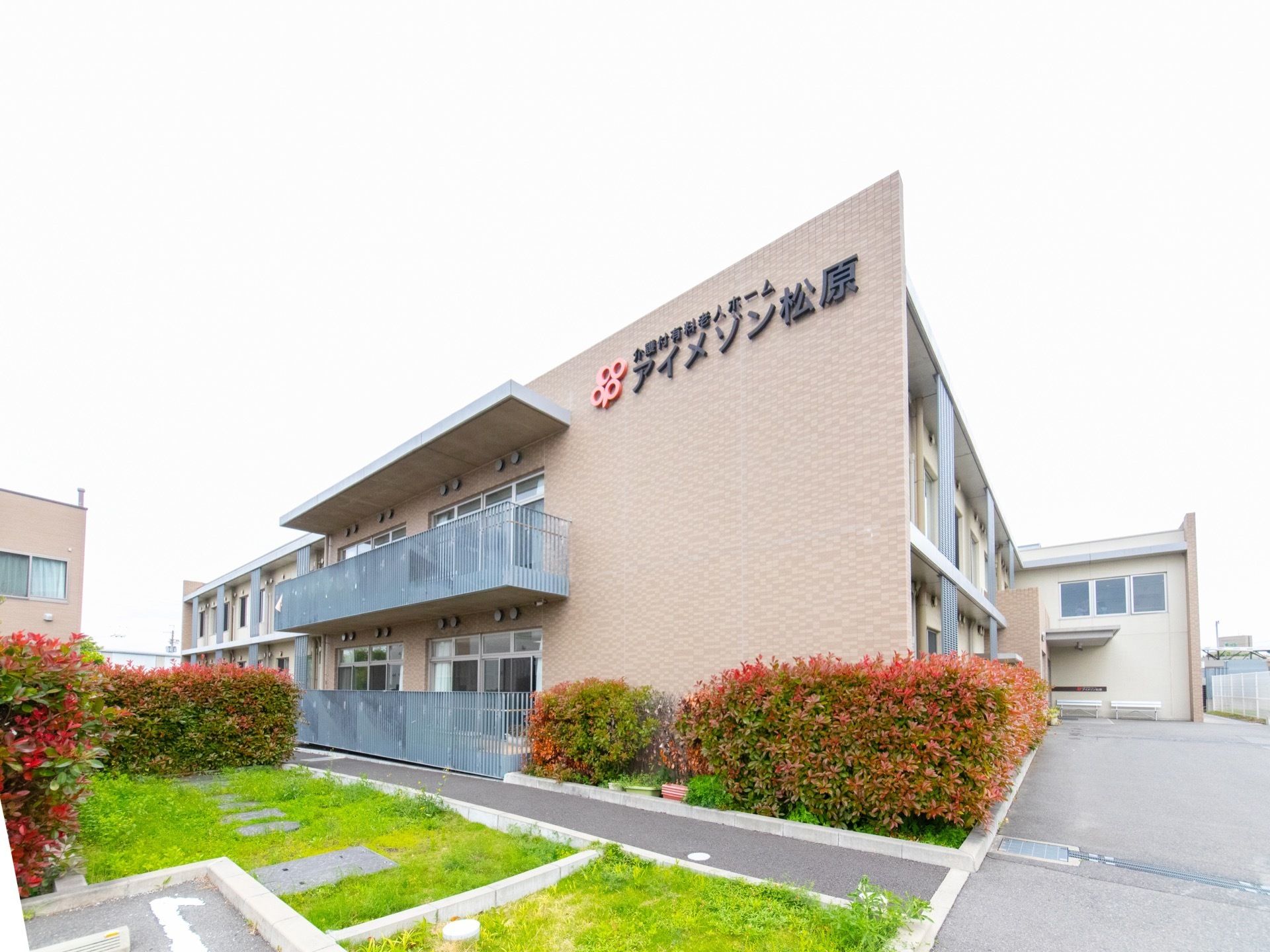 建物は２階建てで、壁面は全体がピンク色に塗られており、植栽に囲まれている。上部に施設名が大きく表示されている。