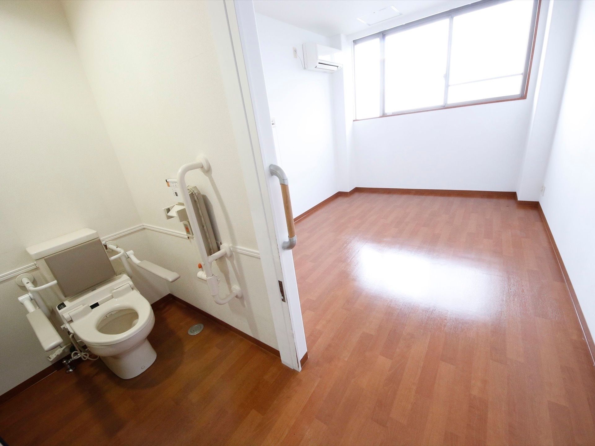 トイレの扉が開いていて、洋式便器が見えている。ベッドは置いておらず、すっきりしている。エアコンが設置されている。
