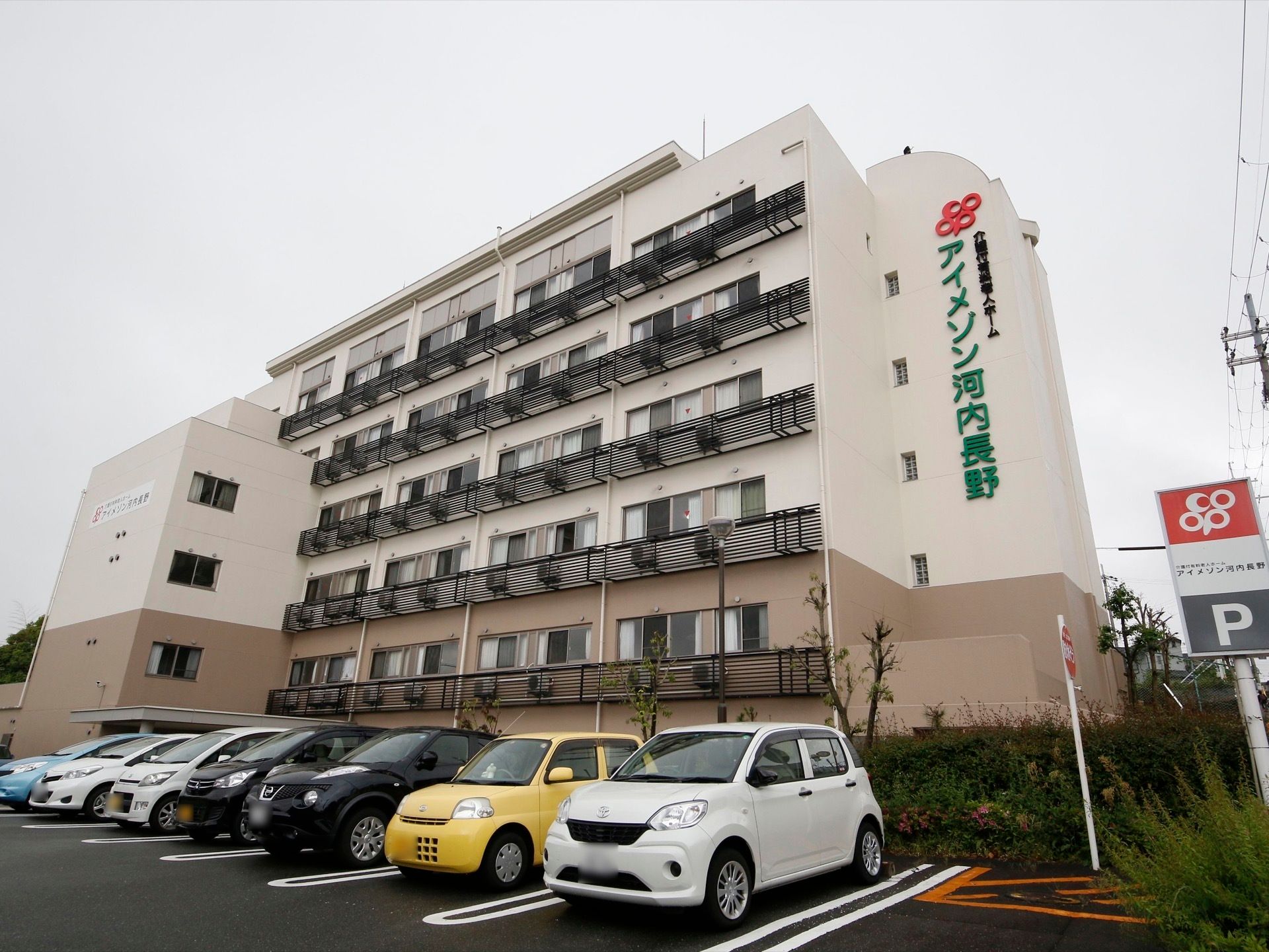 5階建ての建物が見える。各階には居室の窓が見え、エアコンの室外機が設置されている。施設名が大きく掲示されている。