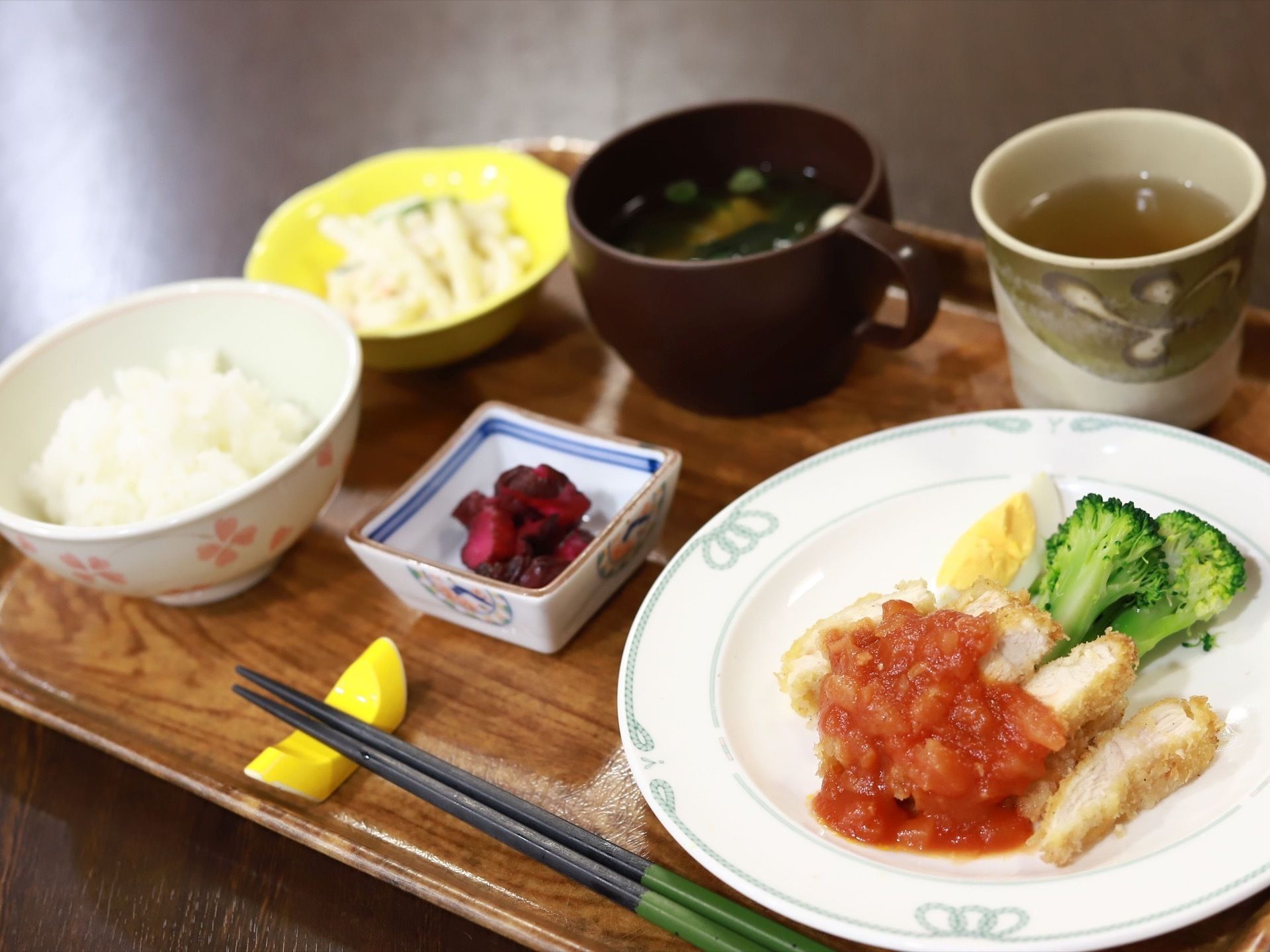 手前の皿にチキンカツ、ゆで卵、ブロッコリーが盛られている。中央にはきゅうりの漬物が入った皿がある。ご飯、味噌汁の他にもう一品用意されている。