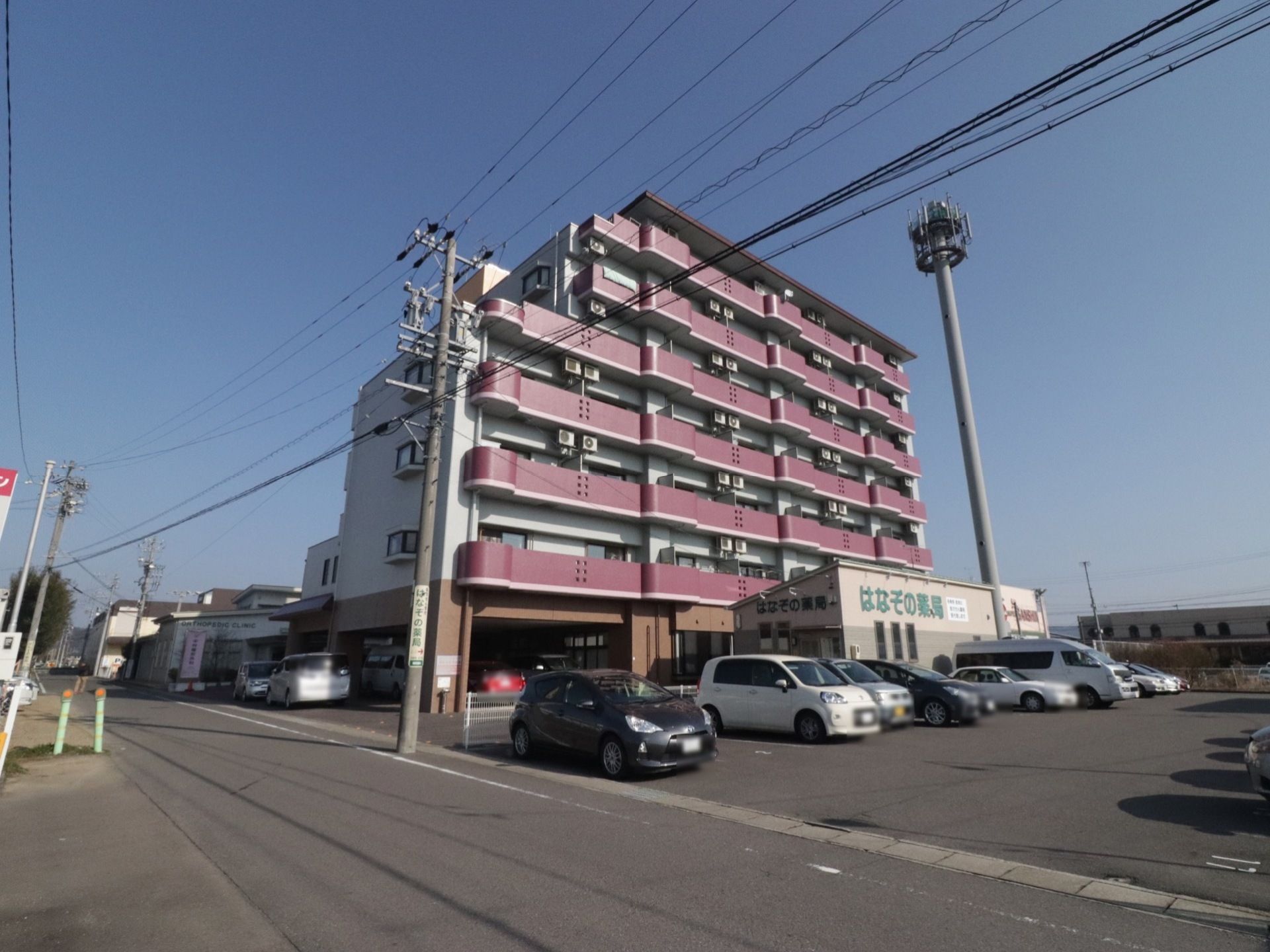 白壁に赤のベランダが明るく目立つ７階建ての施設の様子。大きな駐車場を備えているので、お車で来訪される方には便利である。