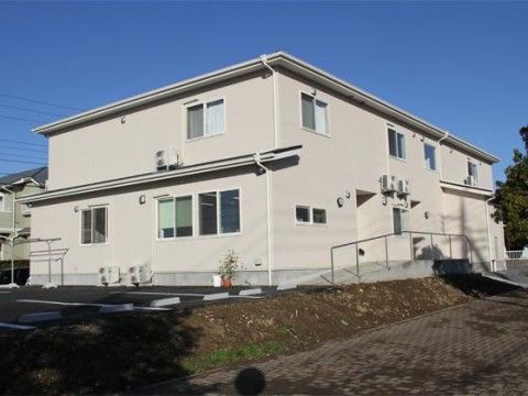 住宅街に馴染むオフホワイトの２階建ての建物。建物の横には駐車場があり、玄関まではスローブとなっている。