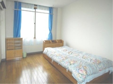 白を基調とした明るい室内。窓にはレースのカーテンがかけられ柔らかい日差しが入る。ベッドとエアコン、タンスが設置されている。